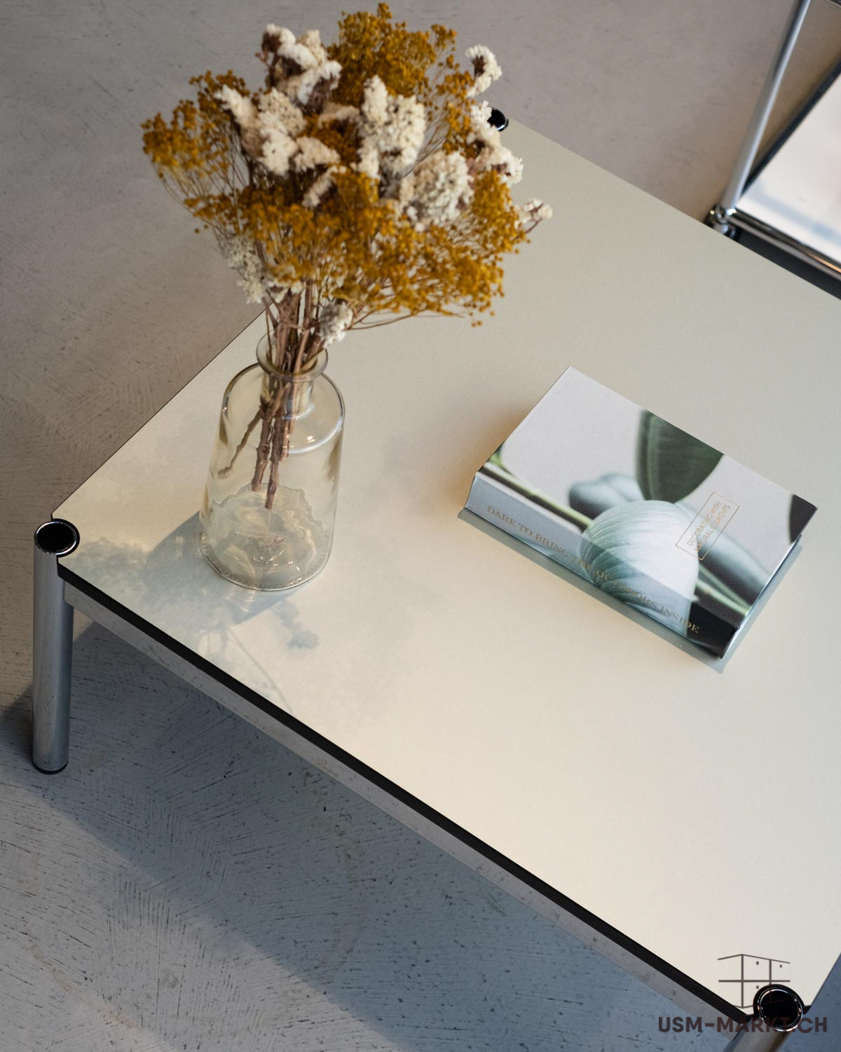 Tavolino minimalista color beige chiaro con gambe cromate. Sopra, un vaso di vetro trasparente con fiori secchi gialli e bianchi. Accanto, un libro con copertina verde, forma rotonda e titolo "DARE TO BRING COLORS INSIDE". Pavimento in cemento in ombra.