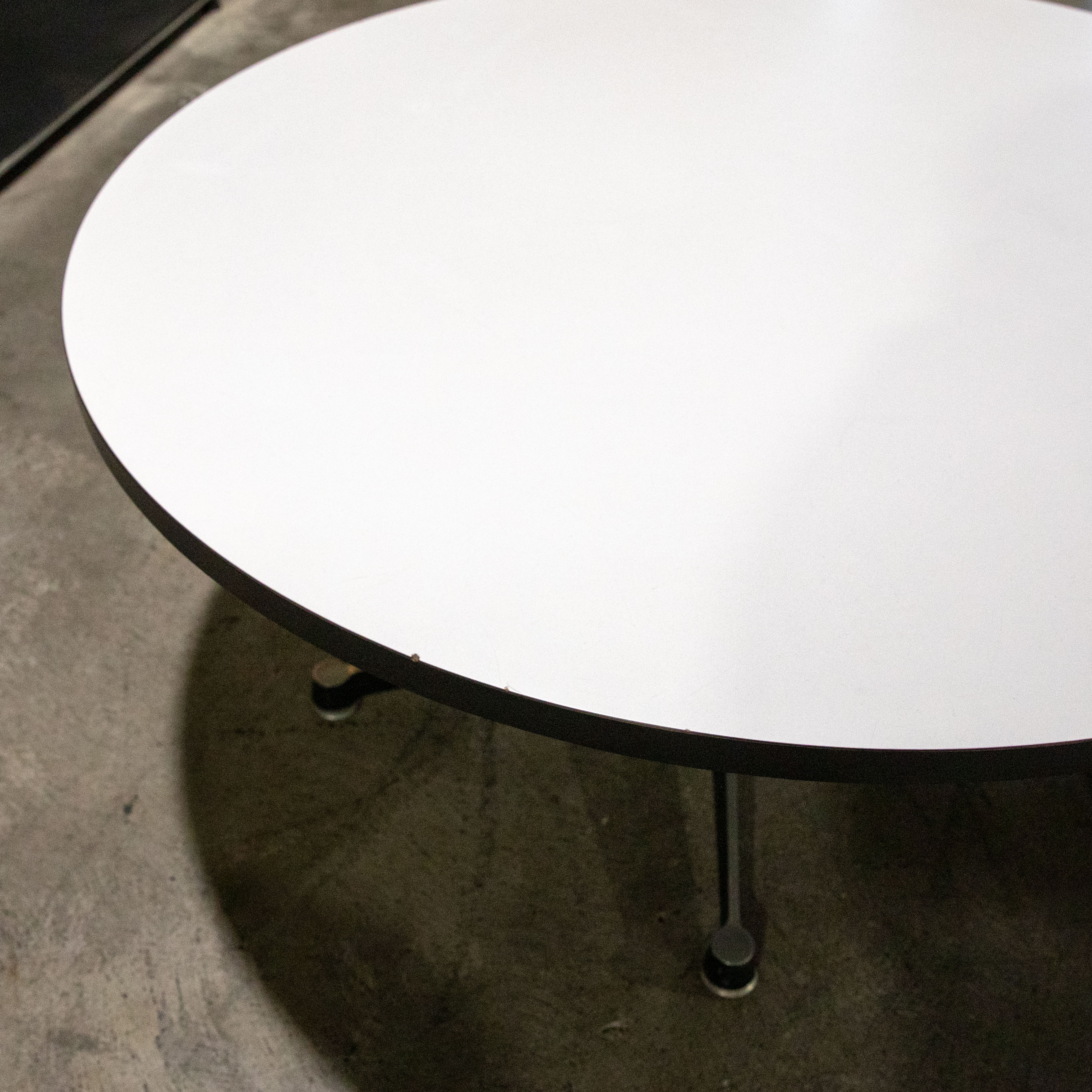 Runder, weißer Charles & Ray Eames Sofatisch von Hermann Miller. Helle Tischplatte mit dunklem Rand, leichten Gebrauchsspuren und Abplatzern. Dunkles Metallbein unten rechts mit Schatten auf grauem Betonboden.