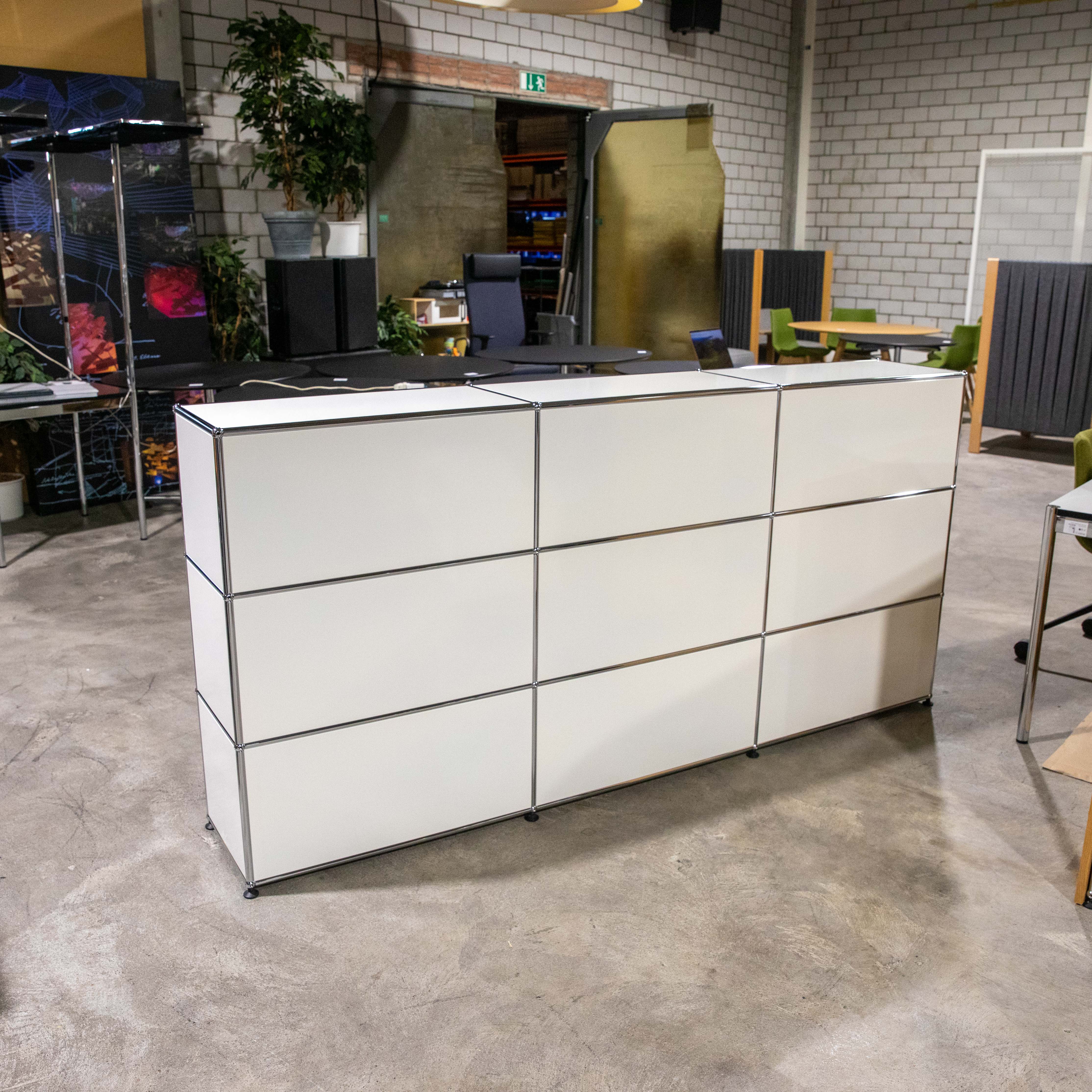 Freistehendes weißes USM Haller Regal mit drei Reihen und drei Spalten (neun offene Fächer). Klare weiße Platten und glänzende Chromverbindungen zeigen das modulare Design. Auf hellgrauem Betonboden in modernem Raum mit Ziegelwänden.