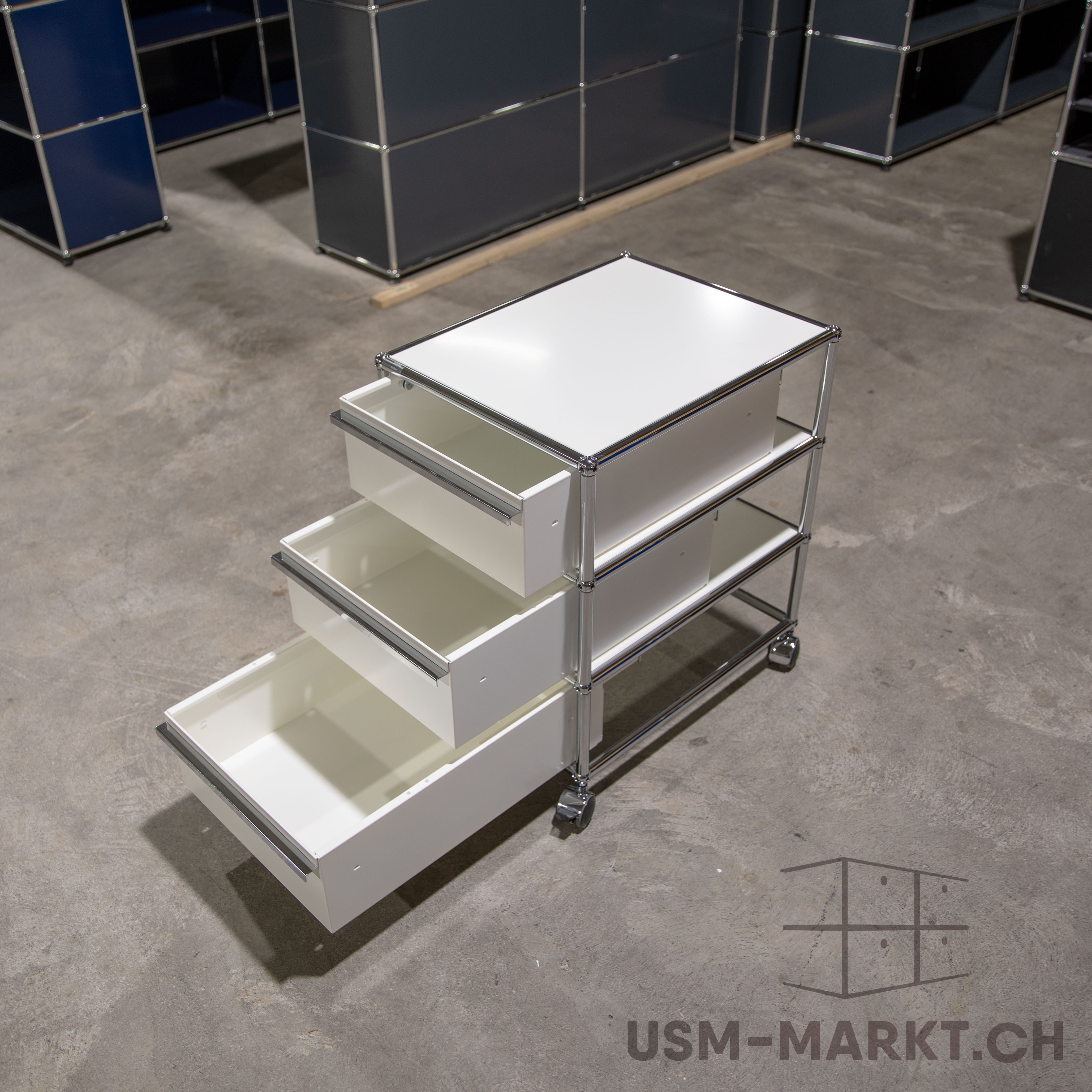 Carrello USM Haller bianco a tre ripiani con tre cassetti leggermente aperti su un pavimento in cemento grigio. Struttura cromata lucida con piccole rotelle. Sfocati sullo sfondo: altri mobili modulari nei toni del blu scuro e del grigio.