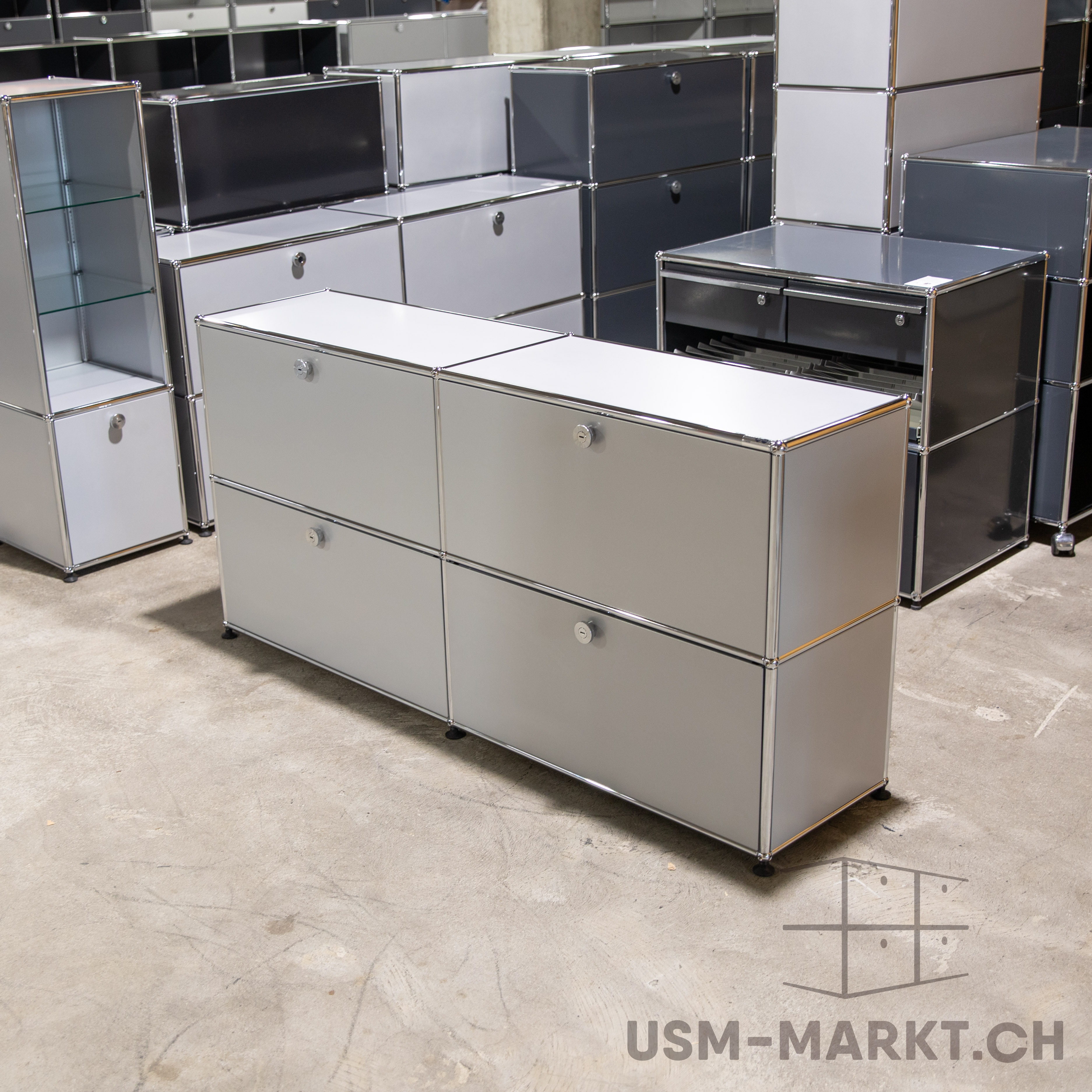 Helles, langes USM Haller Sideboard mit vier geschlossenen Fächern. Dahinter weitere modulare USM Haller Möbel in Hell- und Dunkelgrau, darunter ein hohes Regal mit Glastüren und Kommoden mit Schubladen auf grauem Betonboden.