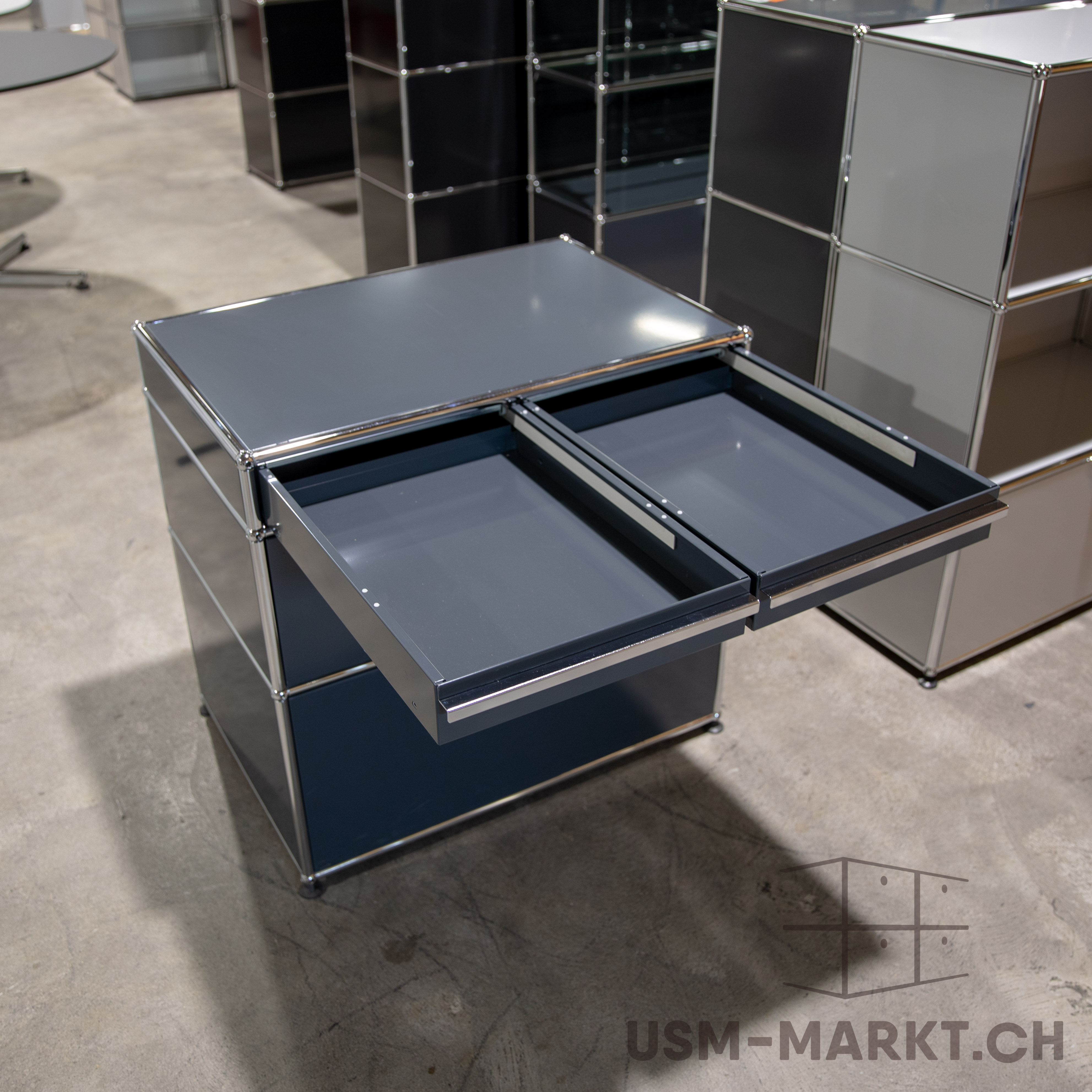 Dunkelgraue USM Haller Kommode mit zwei geöffneten Schubladen, mattgrauen Fronten und Seiten. Modulare Struktur aus glänzendem Chrom. Auf hellgrauem Betonboden; im Hintergrund ähnliche Schränke unscharf. Fokus auf den Schubladen.