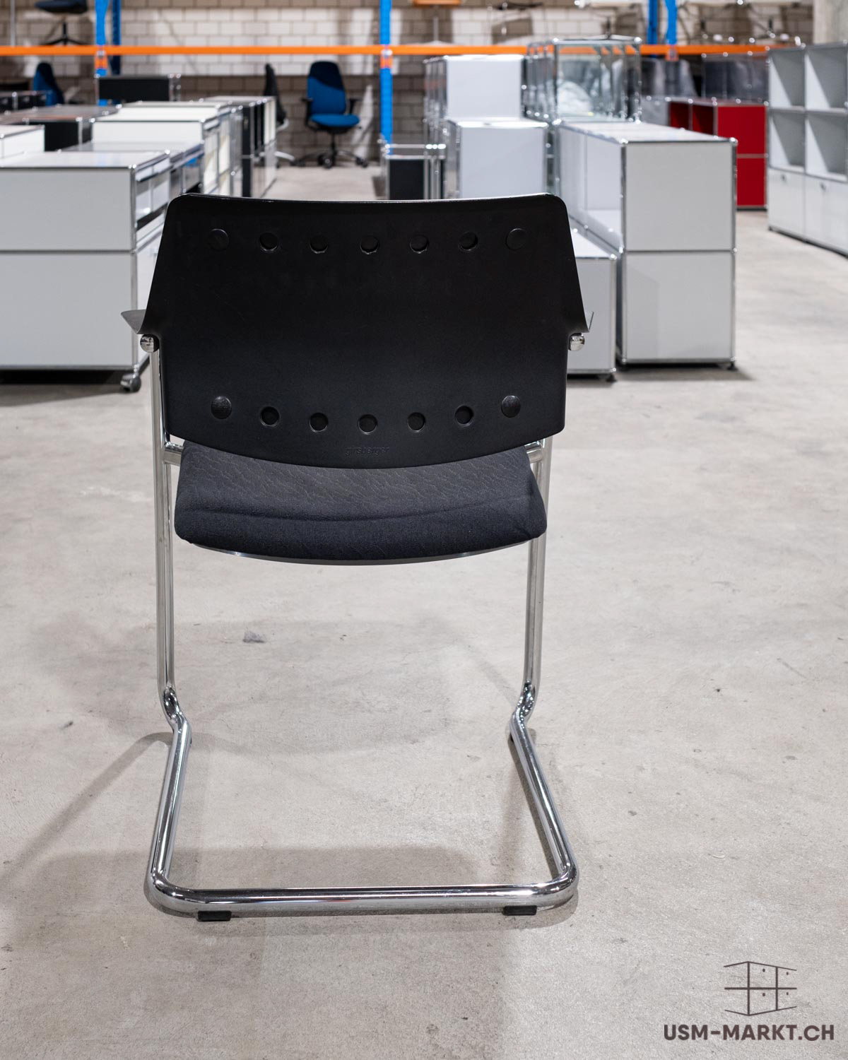 Ein schwarzer Freischwinger-Bürostuhl mit verchromtem Metallgestell steht im Vordergrund auf einem Betonboden. Im Hintergrund sind Reihen von weissen modularen Büromöbeln und vereinzelt blaue Bürostühle in einem grossen Lagerraum zu sehen.