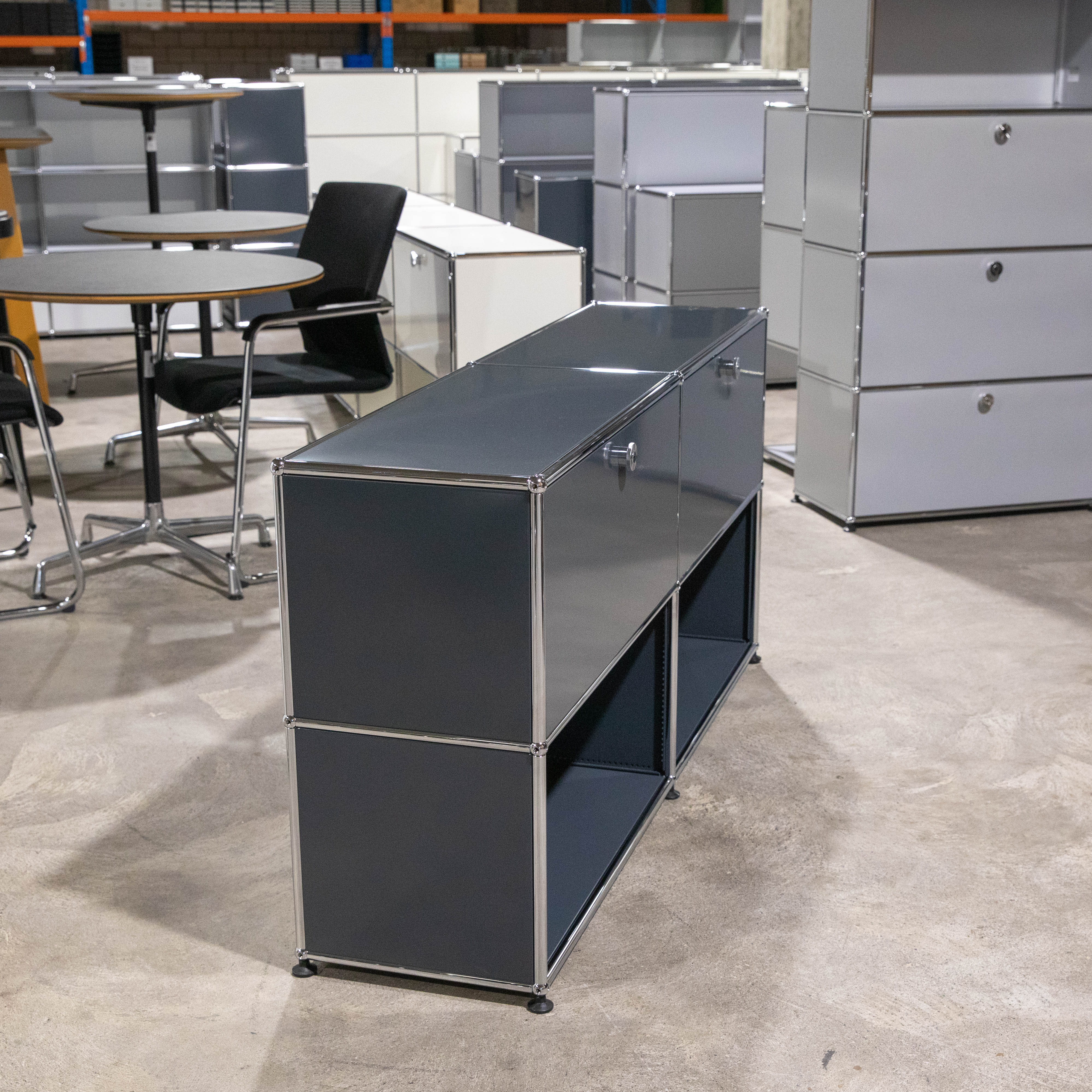 USM Haller Sideboard in Anthrazit, 2 Felder breit und 2 Fächer hoch. Oben klassische Klapptüren mit verchromten Griffen, unten offene Fächer. Das modulare Möbel steht auf hellgrauem Betonboden in einem Lager.