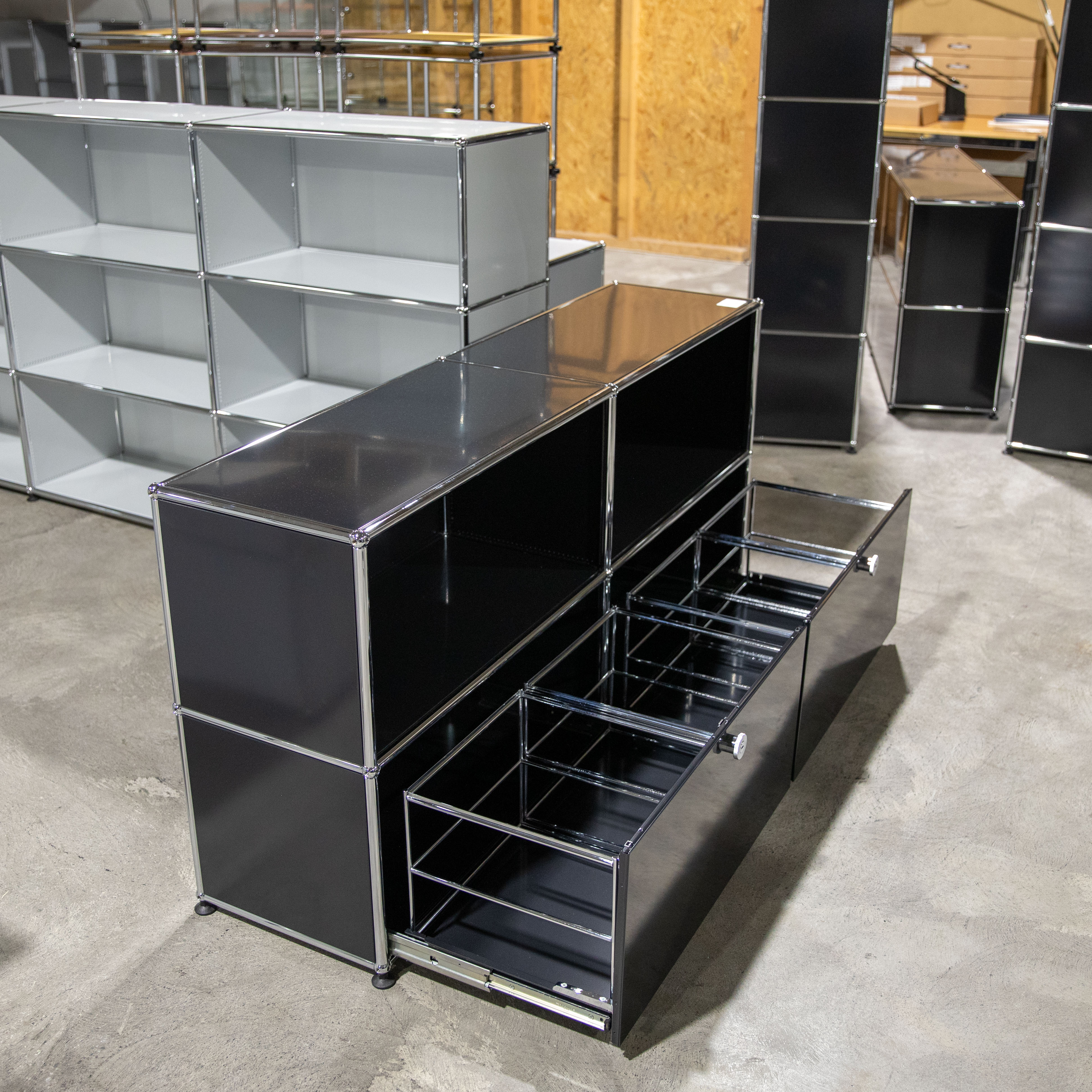 Schwarzes, modulares Sideboard mit glänzender Oberfläche auf Betonboden. Zwei Schubladen mit Metallrahmen für Hängeregister sind geöffnet. Im Hintergrund weitere modulare Regaleinheiten in Hellgrau und Schwarz, eine mit offenen Fächern.