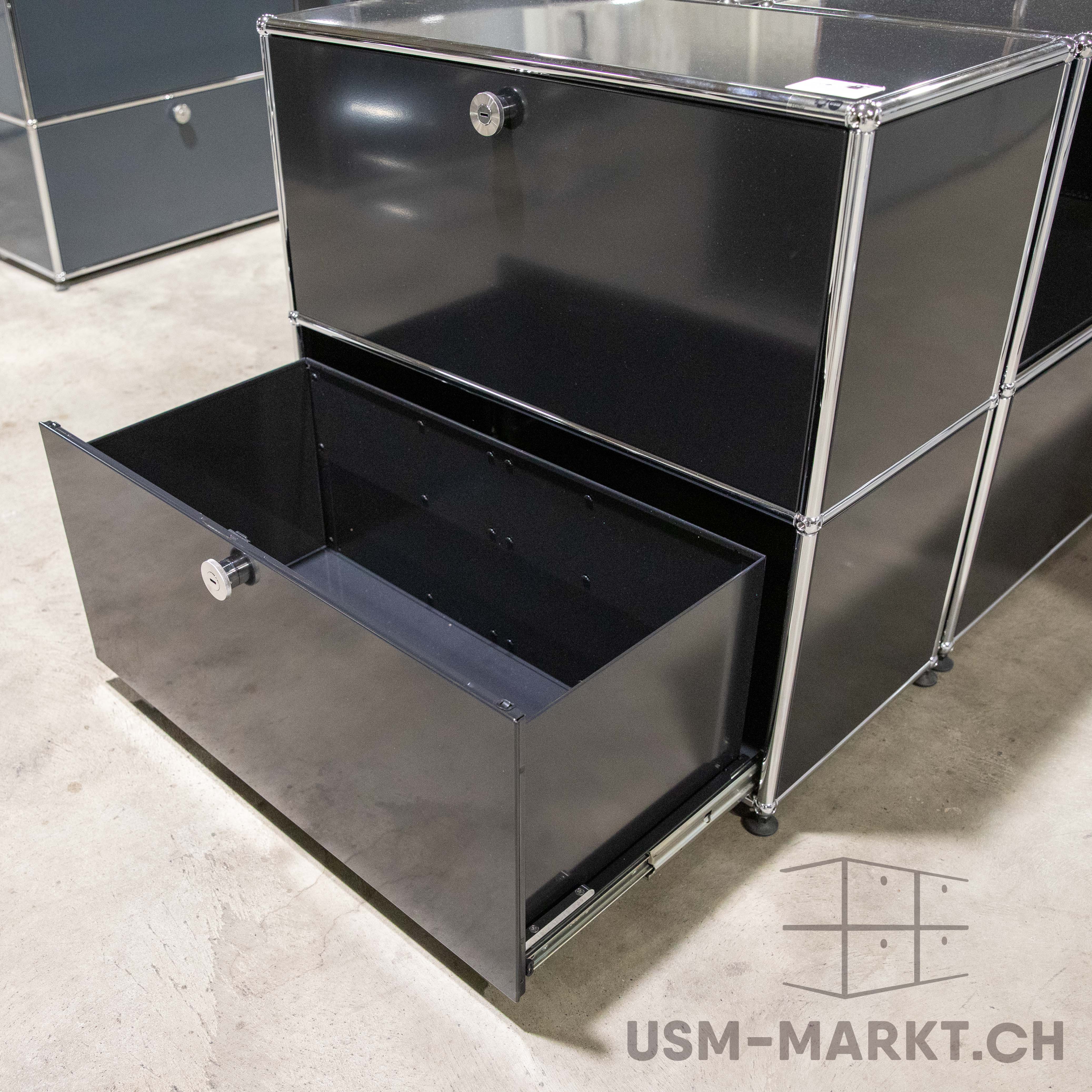 Schwarzes, modulares Schranksystem mit Chromgestell auf grauem Betonboden. Unten links ist ein Schubkasten ganz geöffnet, mit schwarzen Innenwänden und silbernen Auszugsschienen; das Schloss ist sichtbar. Oben ein geschlossener Schubkasten mit Schloss.