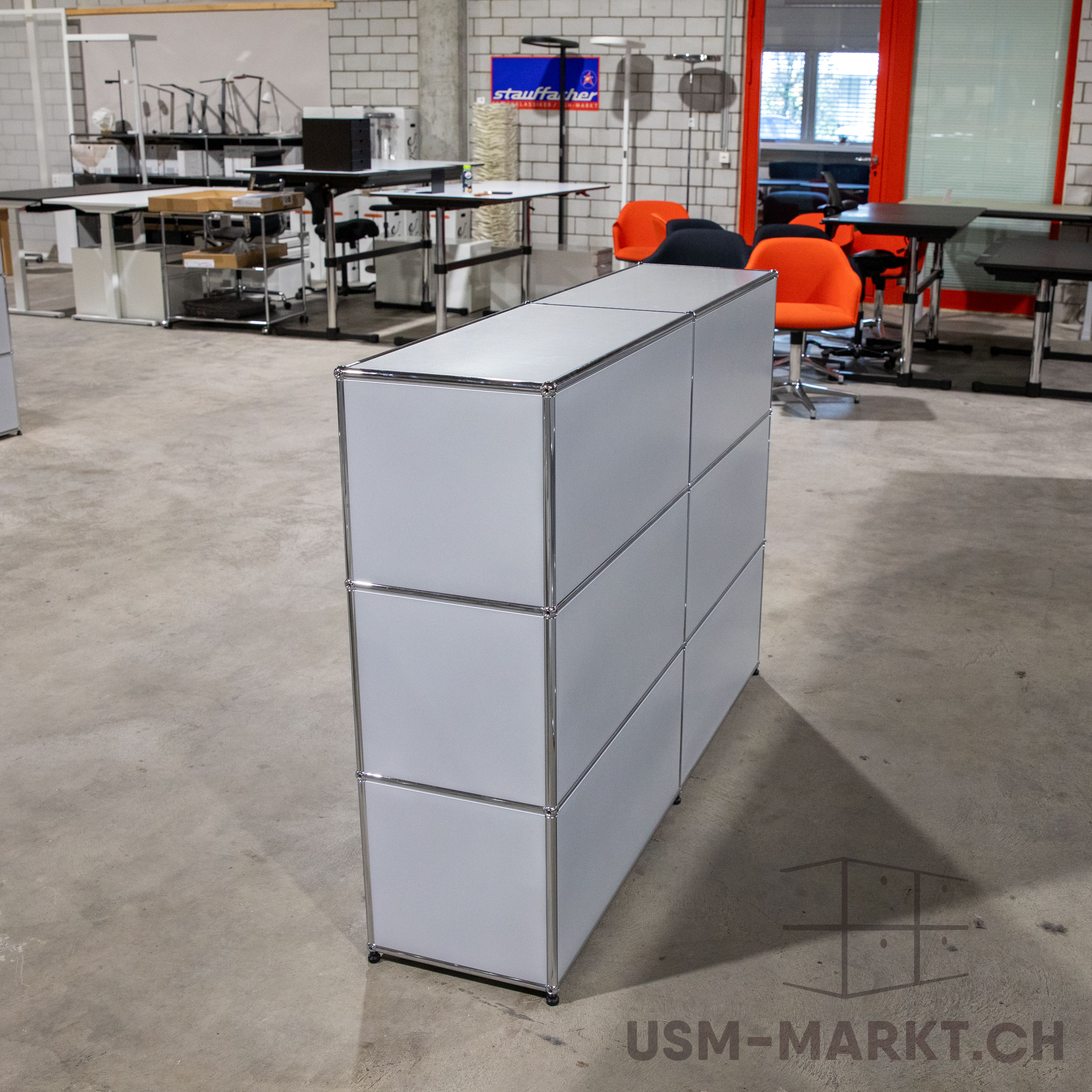 Hellgraues USM Haller Sideboard mit sechs geschlossenen Modulen in zwei Spalten und drei Reihen auf grauem Betonboden. Glänzende Chromrahmen verbinden matte Oberflächen. Im Hintergrund weitere Büromöbel und orange Stühle.