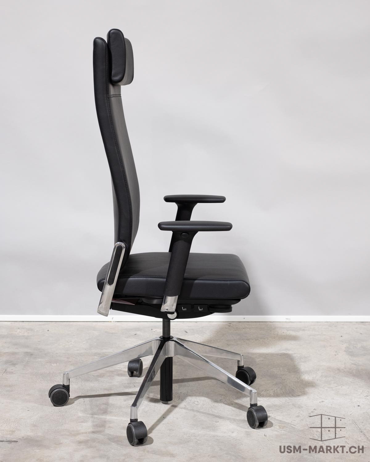 Chaise de bureau moderne noire dotée d’un dossier ergonomique haut et d’un appui-tête rembourré, vue de profil. Accoudoirs noirs, piètement à cinq branches en aluminium poli muni de roulettes, sur un sol en béton gris.
