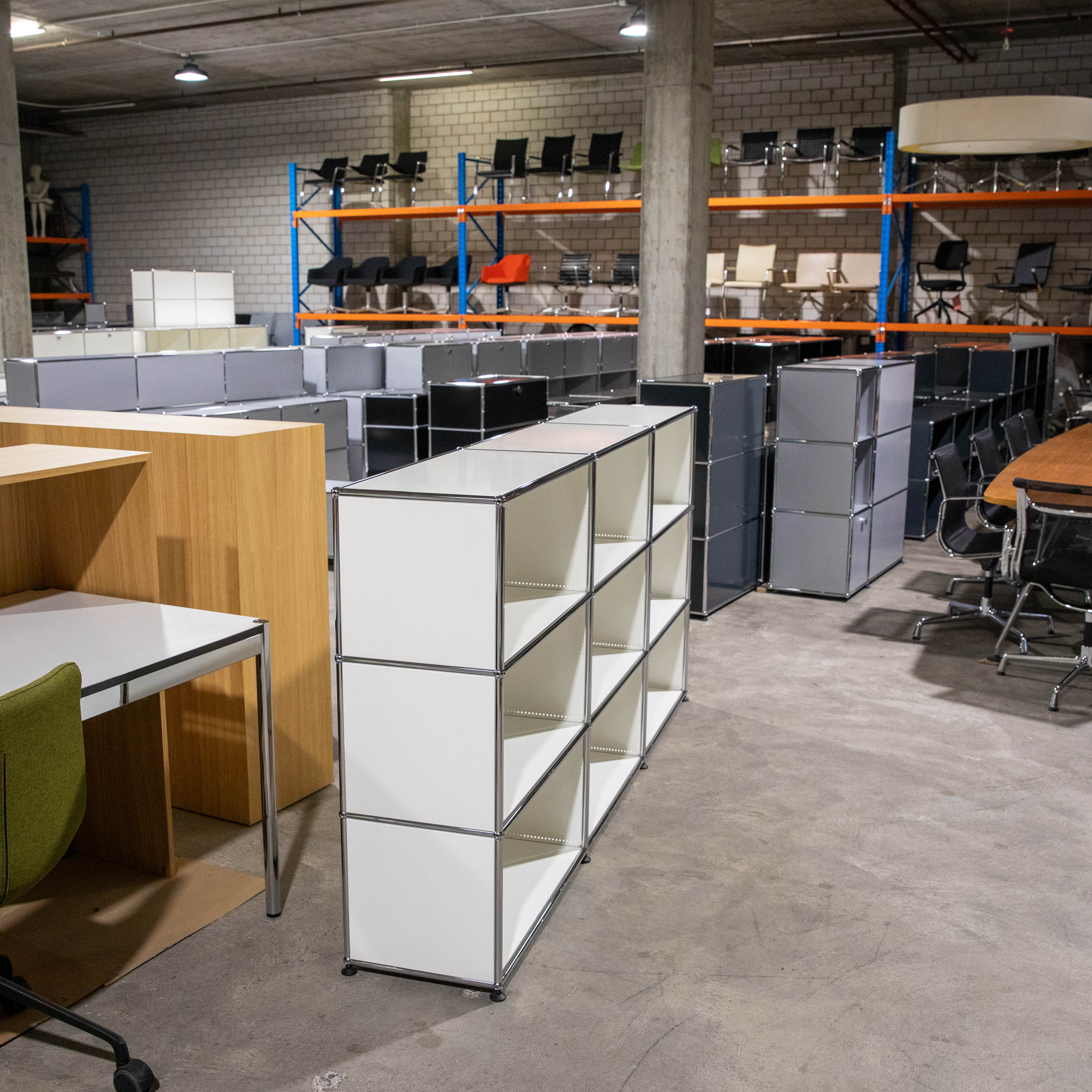Ein gebrauchtes, weisses USM Haller Regal mit drei Reihen und drei Spalten (3x3 Fächer), nach vorne offen, steht auf einem Betonboden. Im Hintergrund sind weitere graue und schwarze USM Haller Möbel sowie Bürostühle in einem grossen Lagerraum zu sehen.