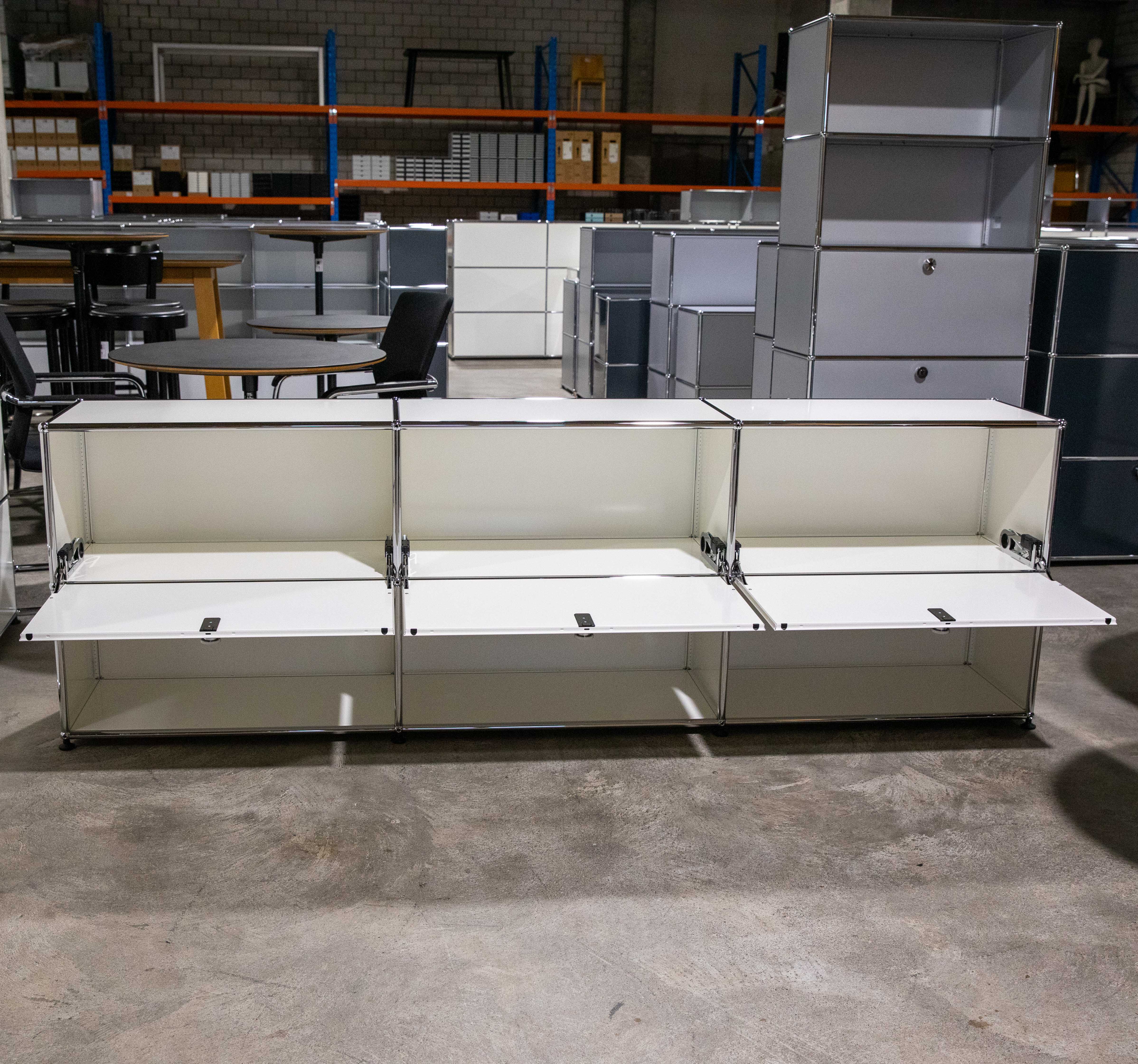 Weißes USM Haller Sideboard mit 3x2 Fächern und drei geöffneten Klapptüren auf grauem Betonboden. Modulares Design aus weißen Metallplatten und Chromrohren, mit sichtbaren unteren Innenfächern. Hintergrund: USM Möbel, Lagerregale.