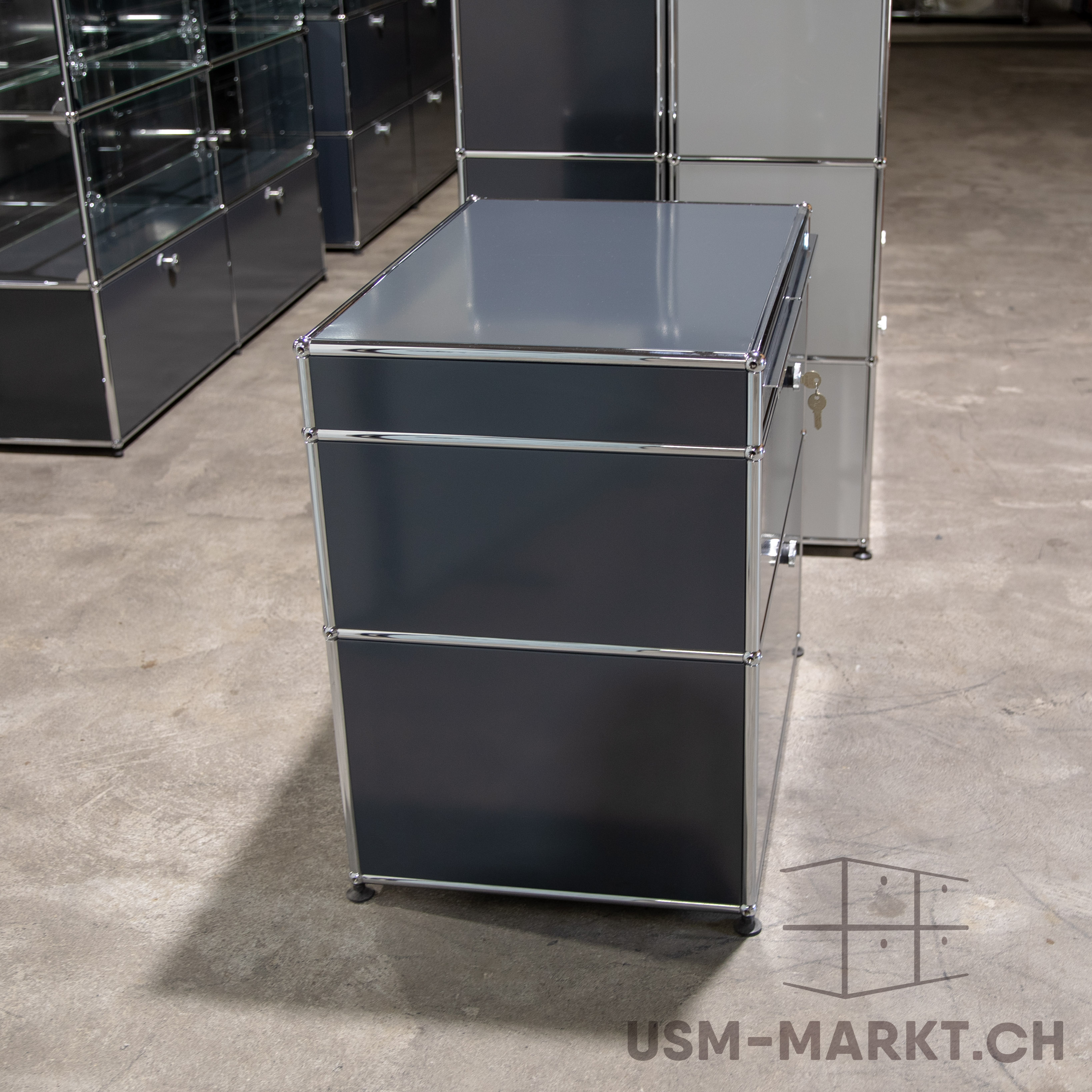 Dreistufiges USM Haller Möbel in Dunkelgrau auf Betonboden. Oben glänzende Oberfläche, darunter zwei matte Schubladen/Türen. Verchromte Rohre verbinden die Elemente. Im Hintergrund weitere USM Möbel. Schlüssel hängt seitlich.