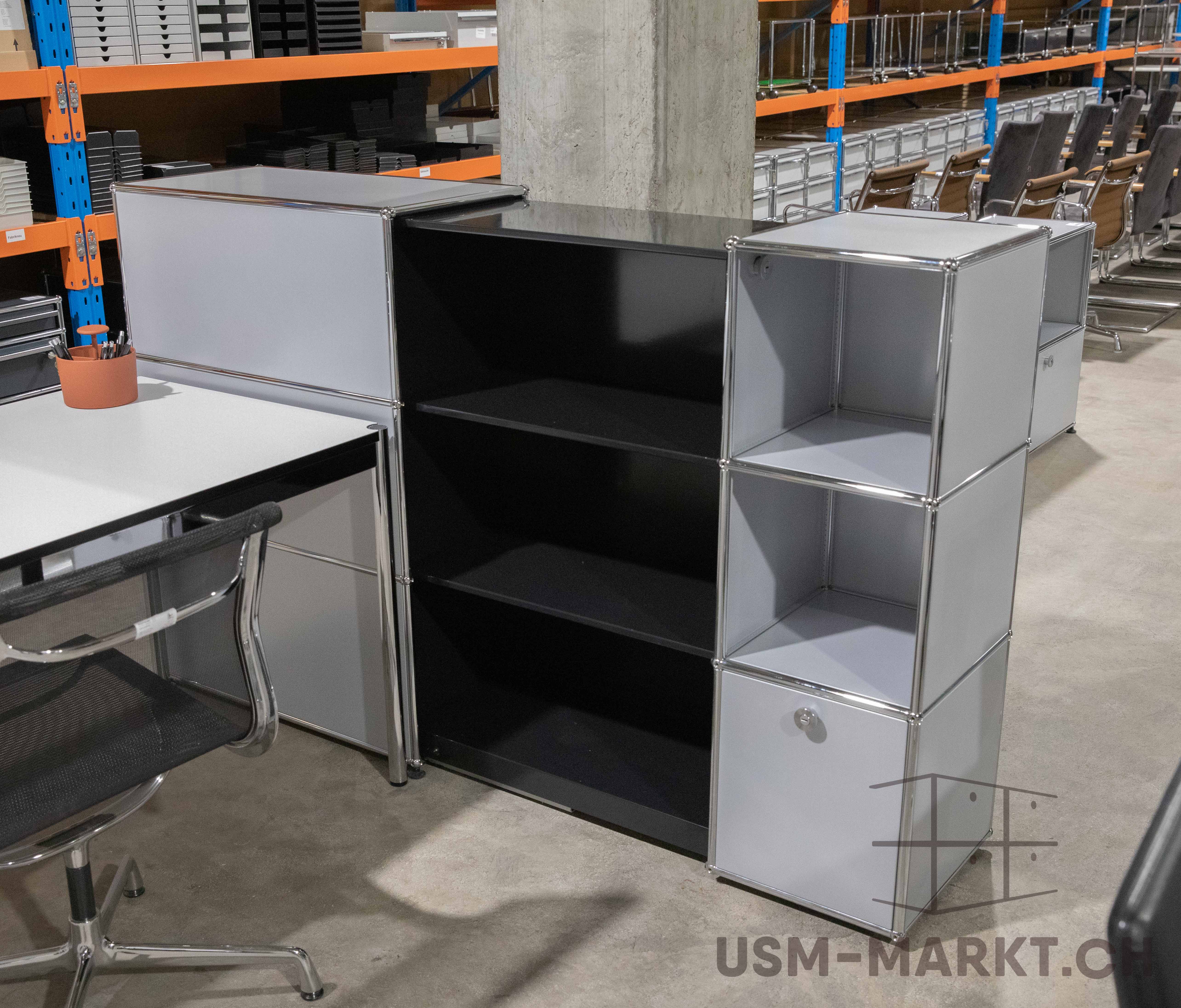 Modernes, modulares Büromöbel-Ensemble in einem Lagerraum: weißer Schreibtisch mit schwarzem Bürostuhl und Regalwand mit hellgrauen geschlossenen sowie offenen schwarzen Fächern. Im Hintergrund weitere Büromöbel in orangenen Lagerregalen.