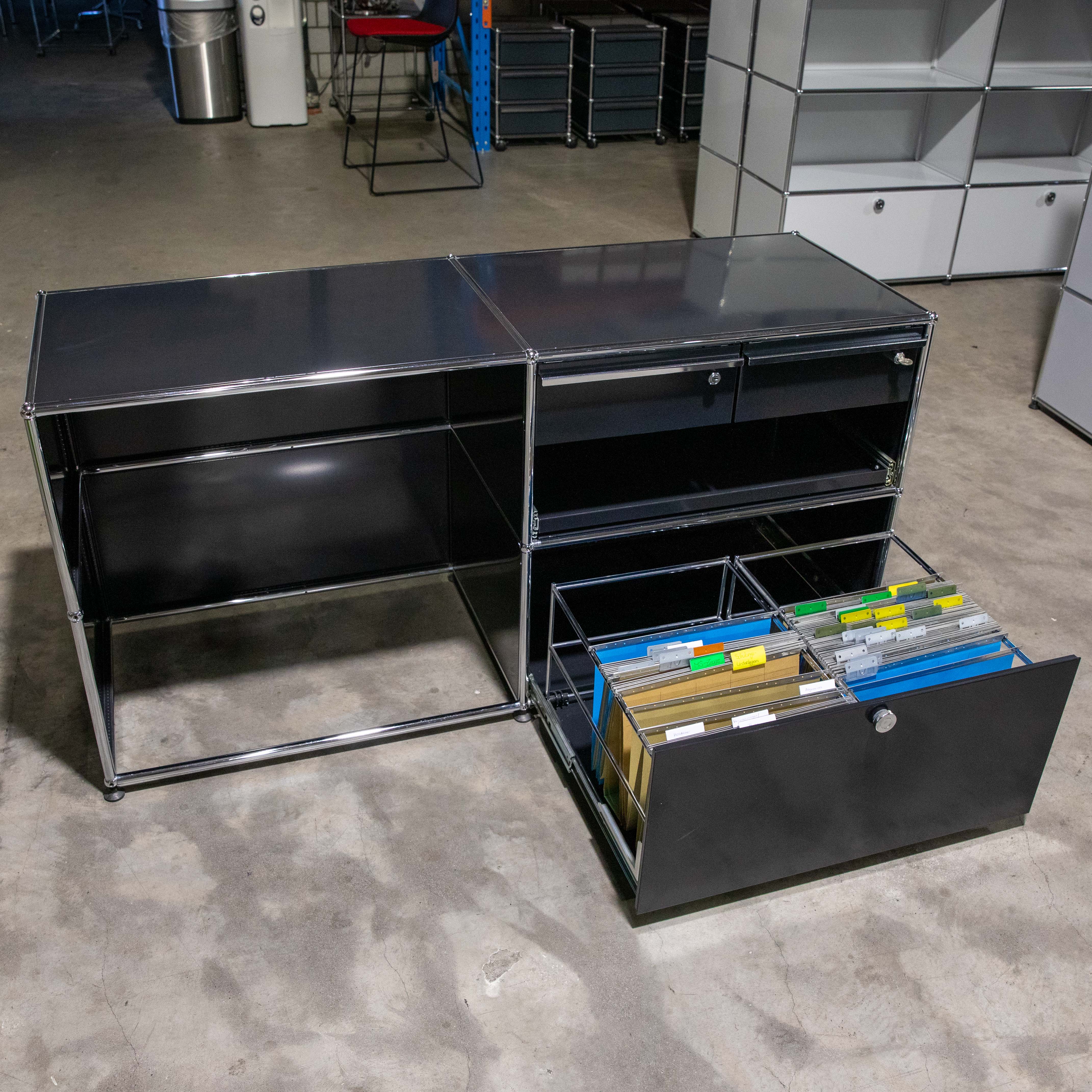Ein großer dunkelgrauer modularer Büroschrank mit Chromrahmen steht auf hellgrauem Betonboden. Der Schrank hat mehrere Fächer und drei Schubladen. Die unterste rechte ist geöffnet mit bunten Hängeregistern in Gelb, Blau, Grün.