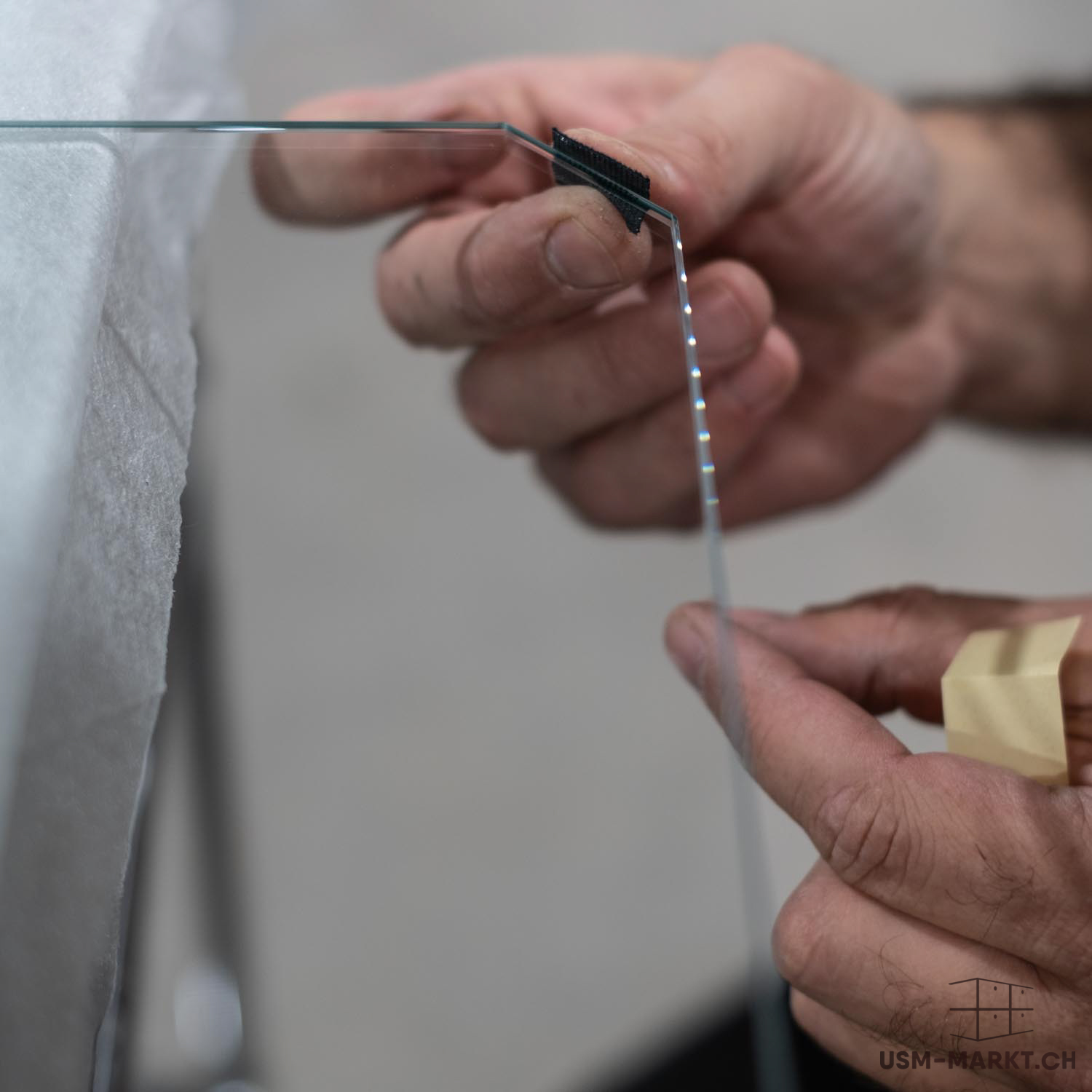 Le mani tengono e piegano con cautela una sottile lastra di vetro, sul cui punto di curvatura è posizionato un morsetto nero. Il bordo del vetro riflette la luce creando punti scintillanti, su uno sfondo bianco e morbido.