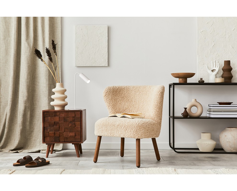 Schlanke, weiße Hansa LED-Tischleuchte auf dunklem Holznachttisch. Daneben beiger Bouclé-Sessel mit offenem Buch. Im Hintergrund heller Vorhang, weiße Strukturwand und schwarzes Regal mit Deko. Modern, gemütlich.