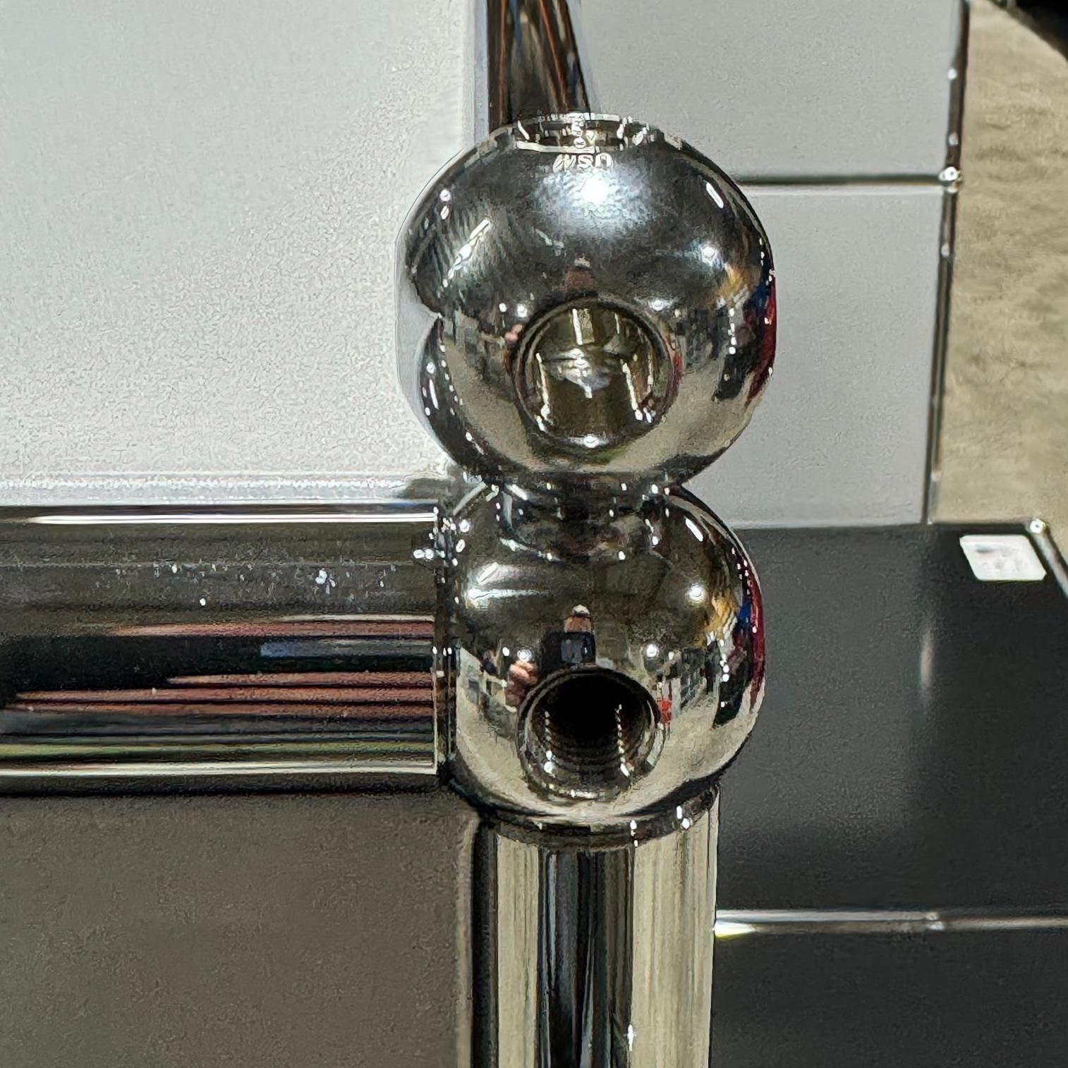 Two shiny chrome ball connectors stacked one on top of the other. The top one has a hexagonal opening and the ‘USM’ logo, whilst the bottom one has a round threaded opening. Connected by a vertical rod in front of blurred light-and-dark panels.