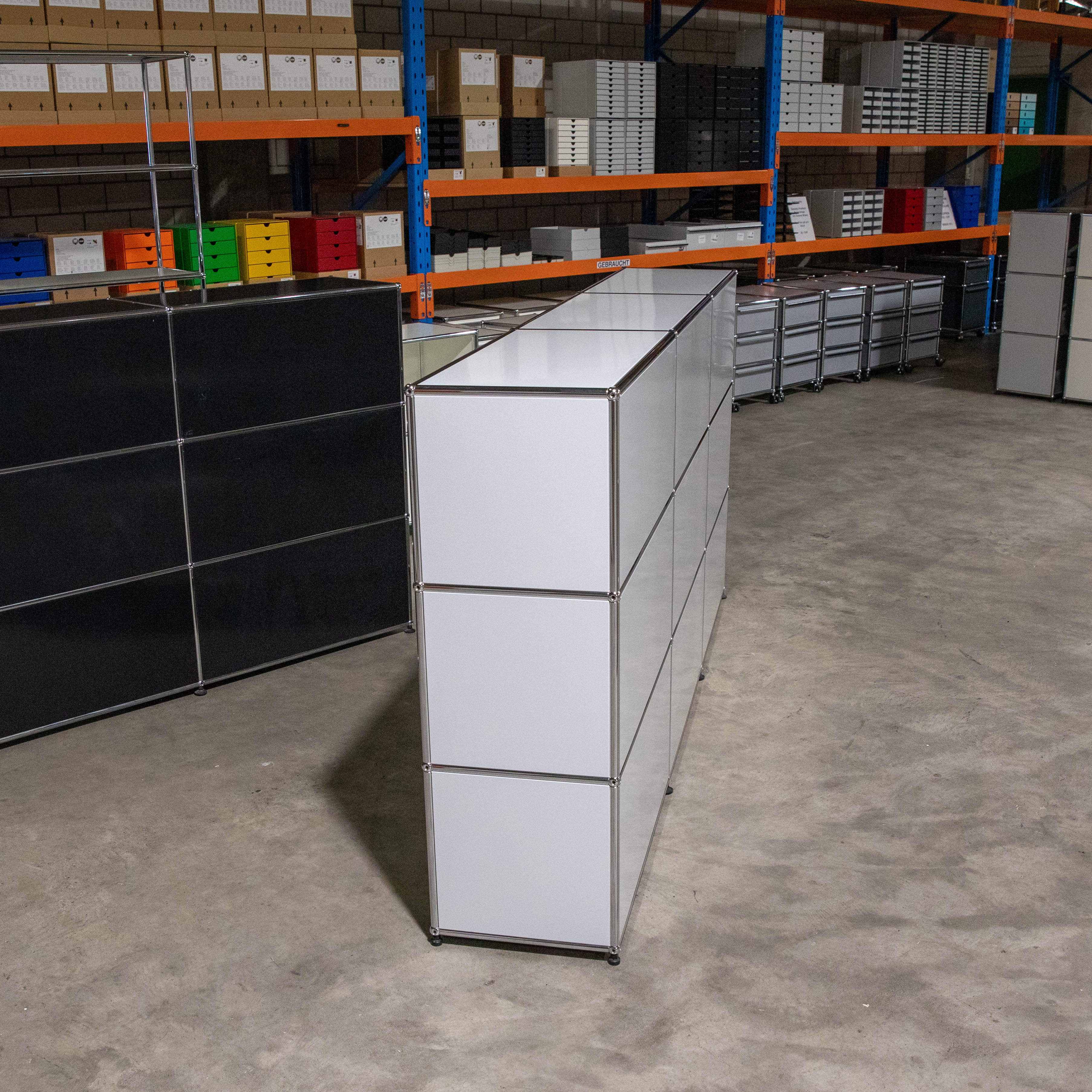 Gebrauchtes USM Haller Regal in Lichtgrau, 3x3-Konfiguration mit neun quadratischen Elementen und modularer Chromstahlrohrkonstruktion. Steht auf Betonboden im Lagerraum, mit weiteren USM Möbeln und Regalen im Hintergrund.