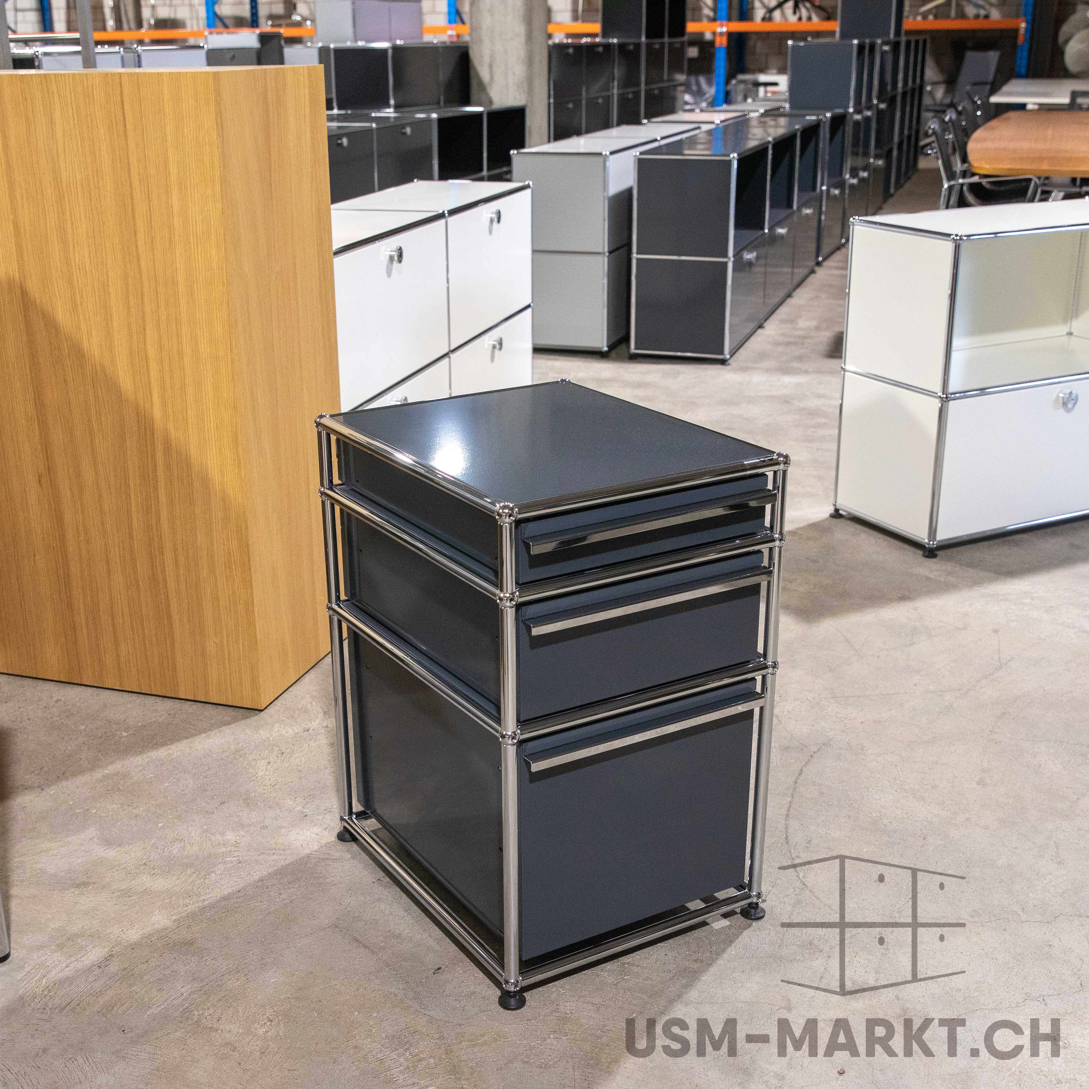 Anthrazitfarbener USM Haller Korpus mit drei Schubladen, umrahmt von glänzenden Chromrohren und schmalen silbernen Griffen. Steht auf grauem Betonboden in hellem Lagerraum, umgeben von USM Möbeln und Holztisch.