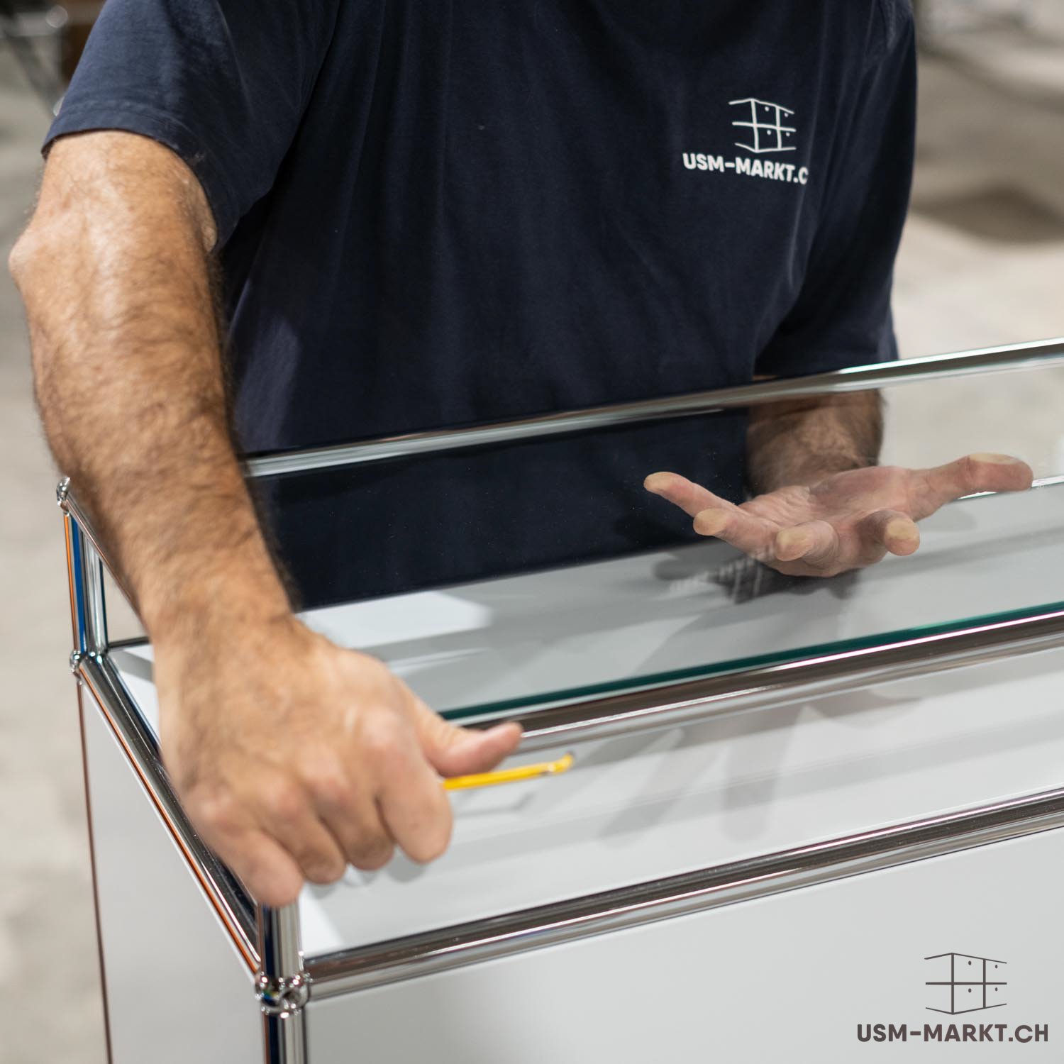 Un uomo con maglietta blu scuro con logo usm-markt monta un mobile modulare bianco. La mano sinistra tiene una matita gialla su un pannello, mentre la destra è sospesa sopra il vetro. Mobili in tubi cromati e pannelli bianchi.