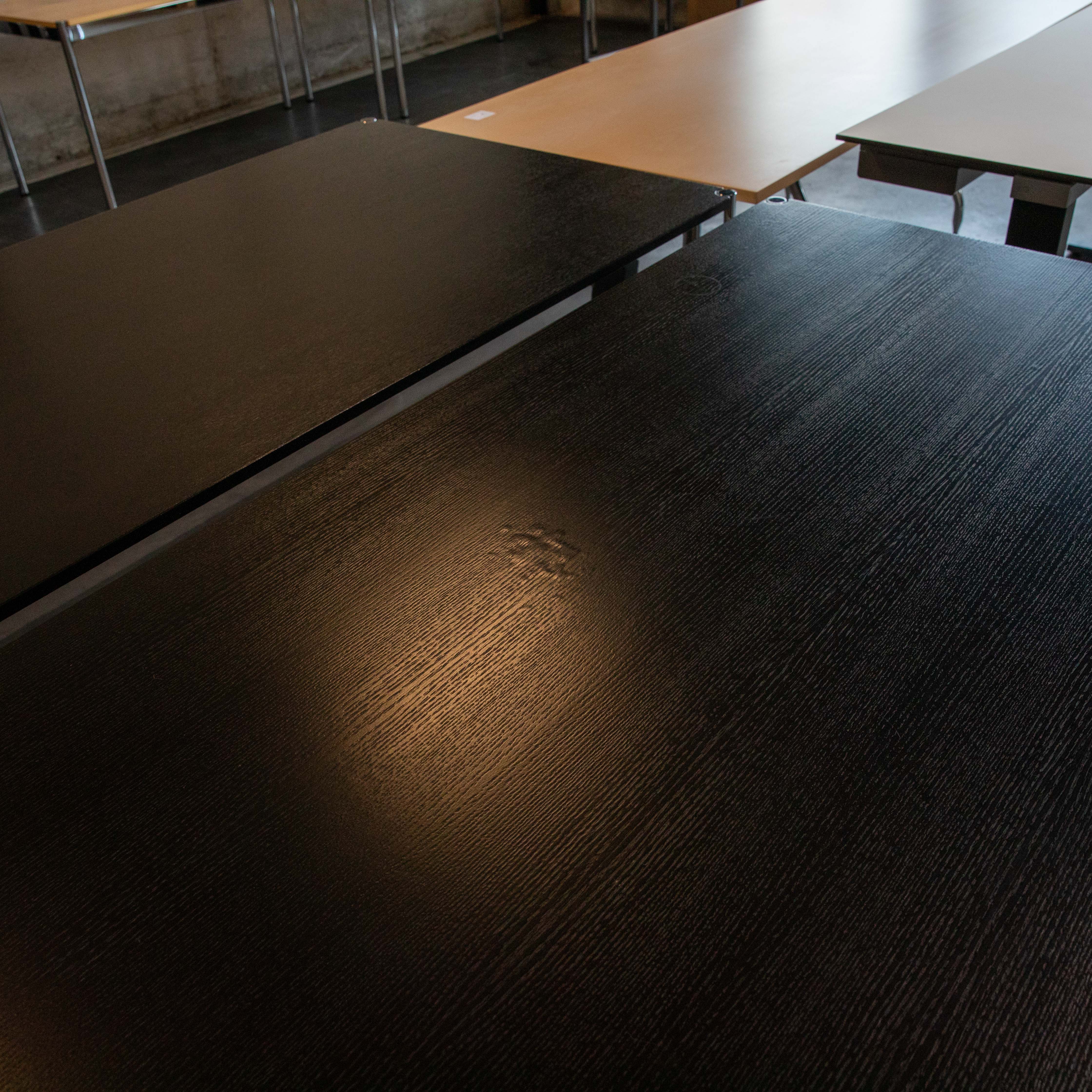 Close-up of the textured, black-lacquered surface of a second-hand USM Haller table made of oak. Water damage, with blistering and slight discolouration, is visible in the centre of the tabletop. Other tables are faintly visible in the background.