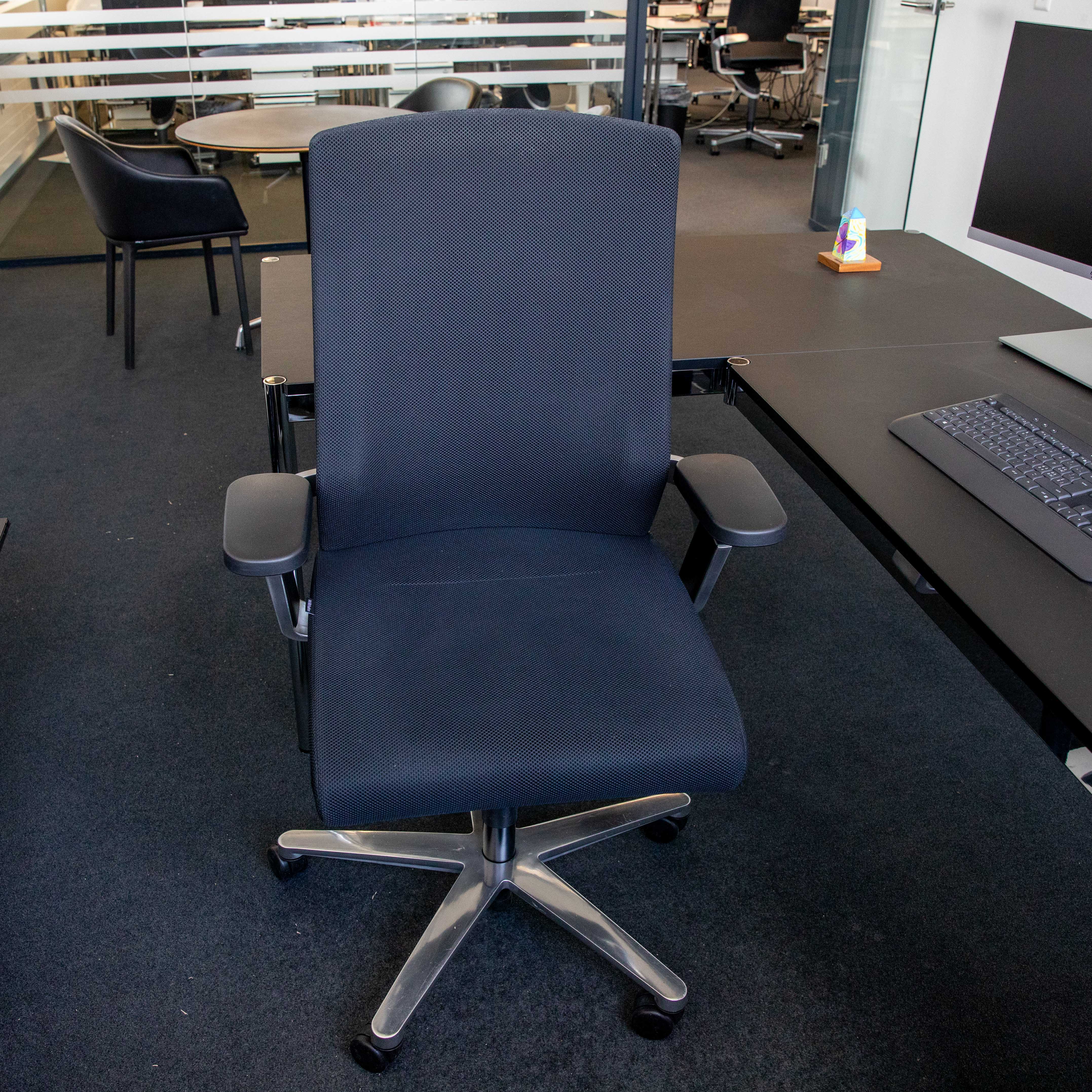 Gebrauchter schwarzer Wilkhahn ON Bürodrehstuhl frontal im Büro. Ergonomische Rückenlehne und Sitzfläche aus schwarzem Netzstoff. Schwarze Armlehnen mit Gebrauchsspuren. Fußkreuz aus poliertem Aluminium mit 5 Rollen.