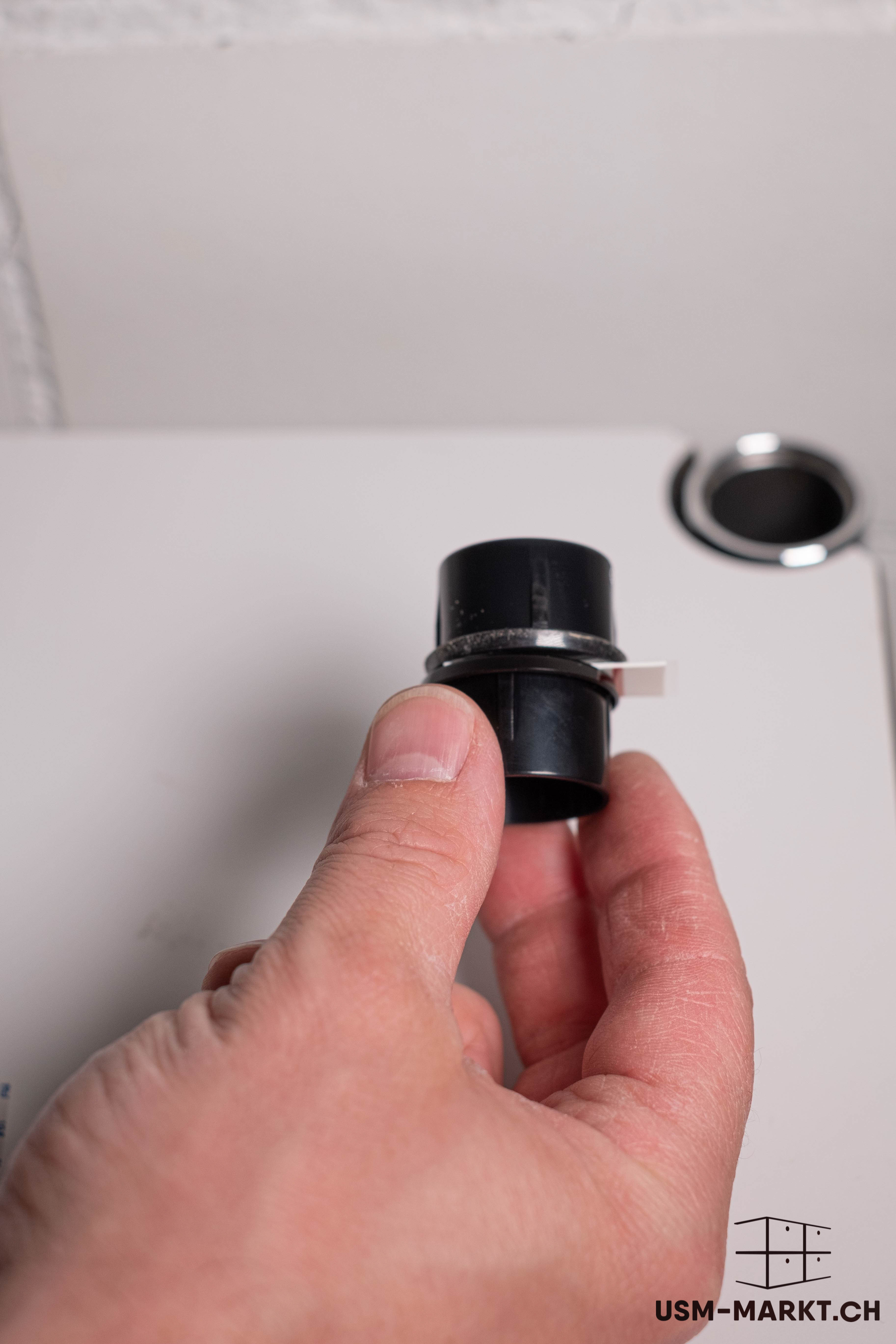 A hand is holding a black, cylindrical plastic connector with a thin white clip, similar to a USM Haller furniture component. Close-up of the smooth component against a blurred light grey background.