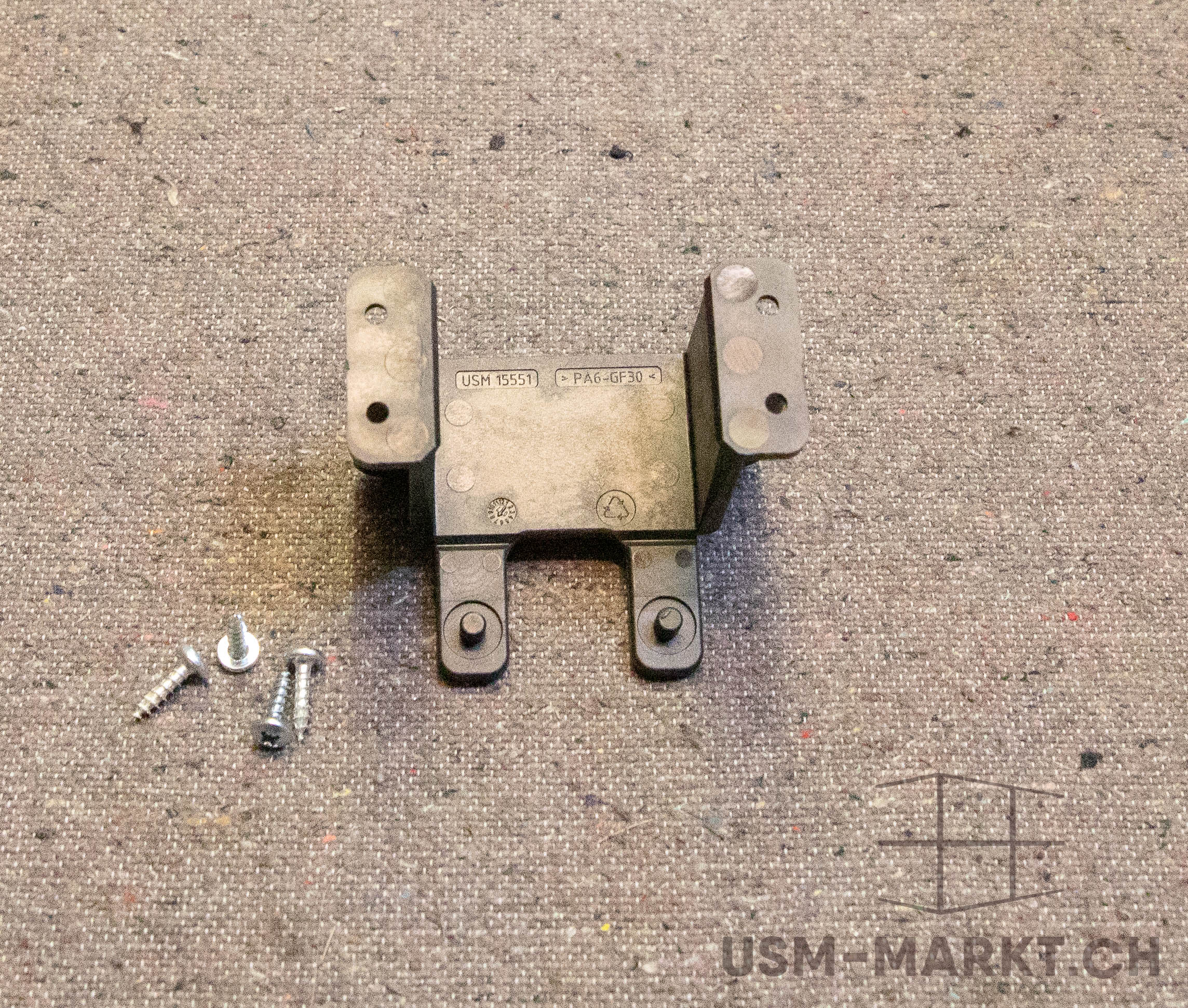 Dark grey plastic bracket with four fixing points and the embossed markings ‘USM 15551’ and ‘PA6-GF30’. Next to it are four silver screws of varying lengths on a light brown patterned surface. Two arms at the top, two feet at the bottom.