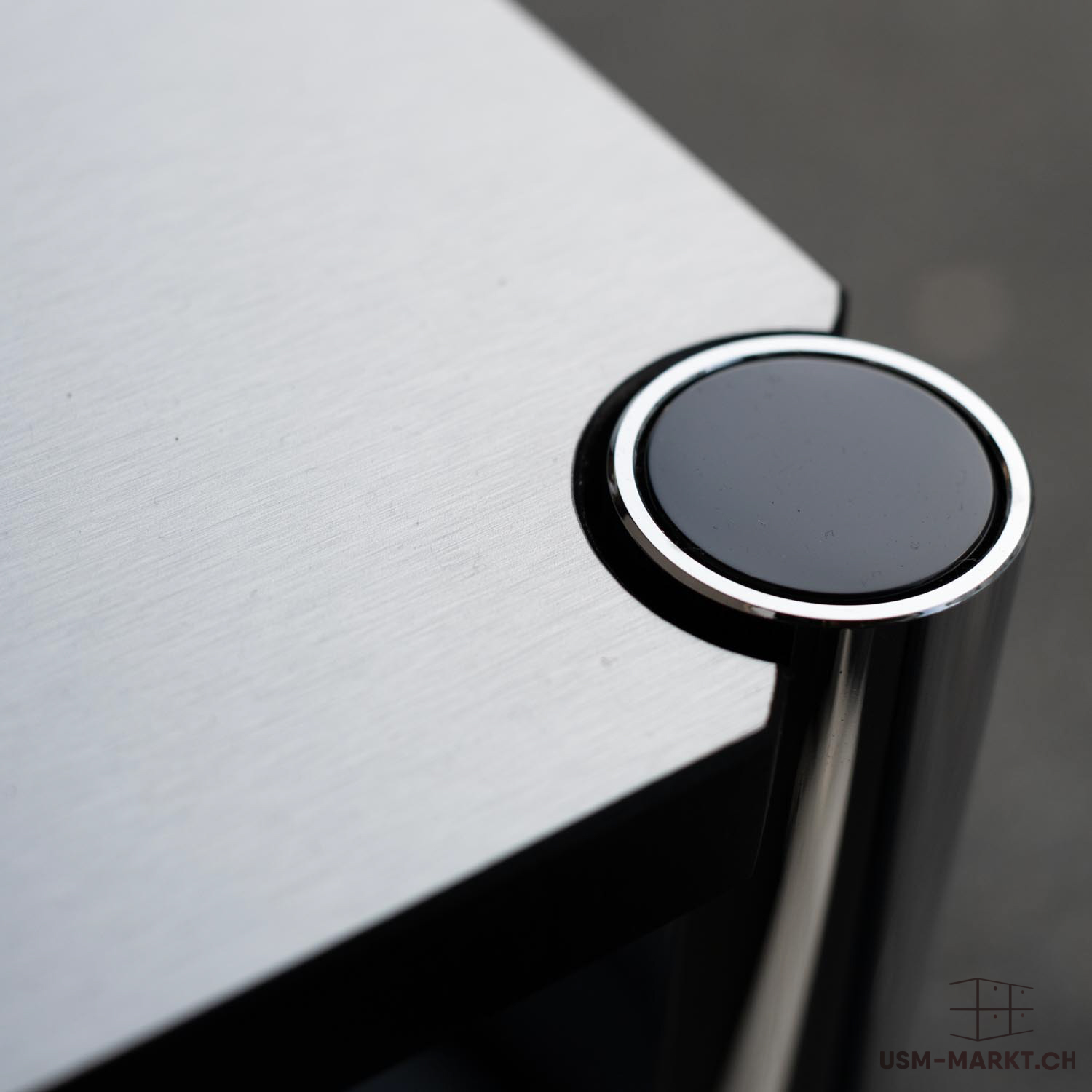 Primo piano dell'angolo di una superficie opaca grigio-argento con sottili striature orizzontali. Questa si unisce a un cappuccio nero lucido di forma rotonda con bordo cromato. Il cappuccio collega la superficie a un montante verticale.
