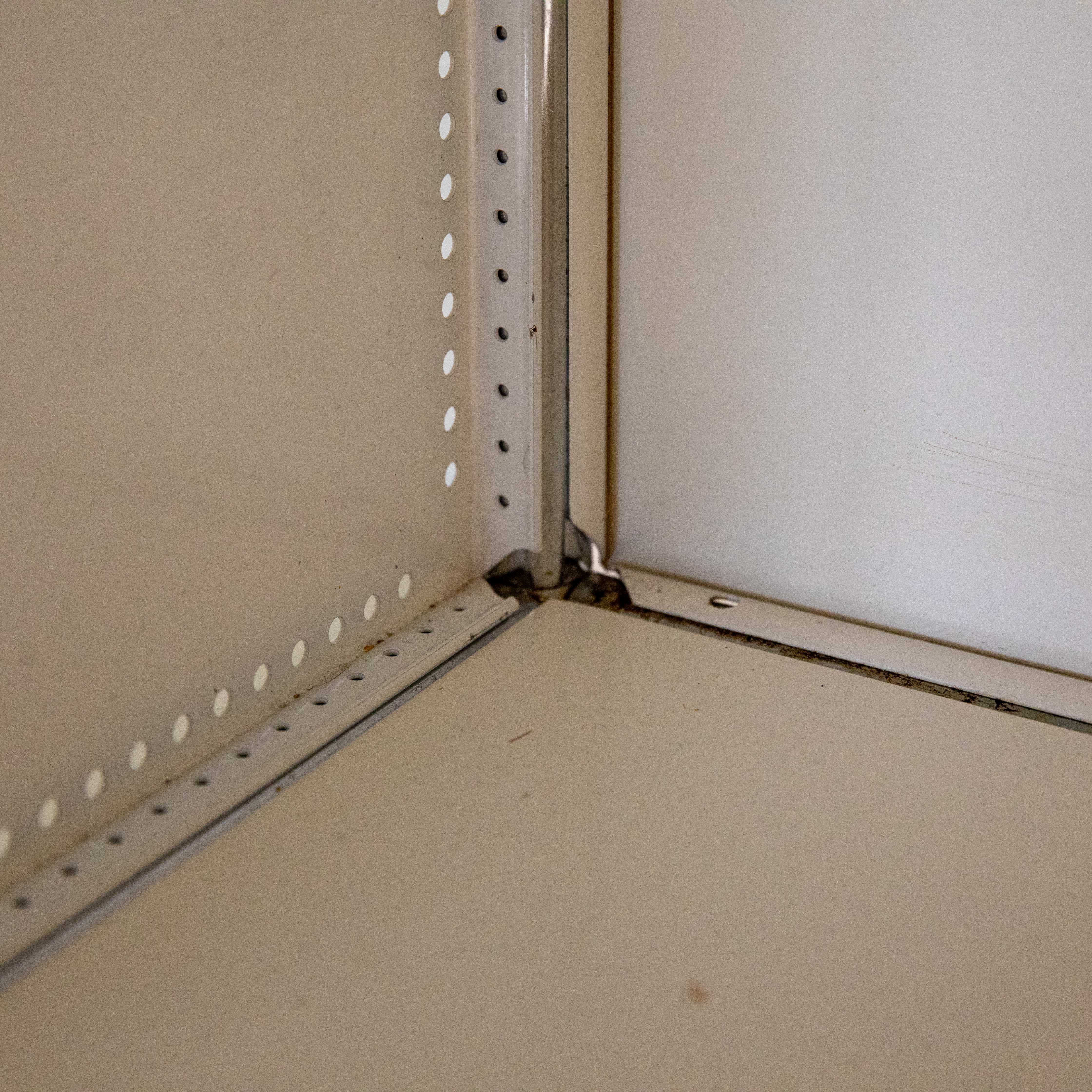 Nahaufnahme der Innenecke eines hellbeigen Metallregals. Vertikale und horizontale Kanten mit gelochten Schienen für Regalböden. Abgenutzte Oberfläche mit Schmutz in der Fuge, besonders in der Ecke. Eindruck von Alter und Gebrauch.