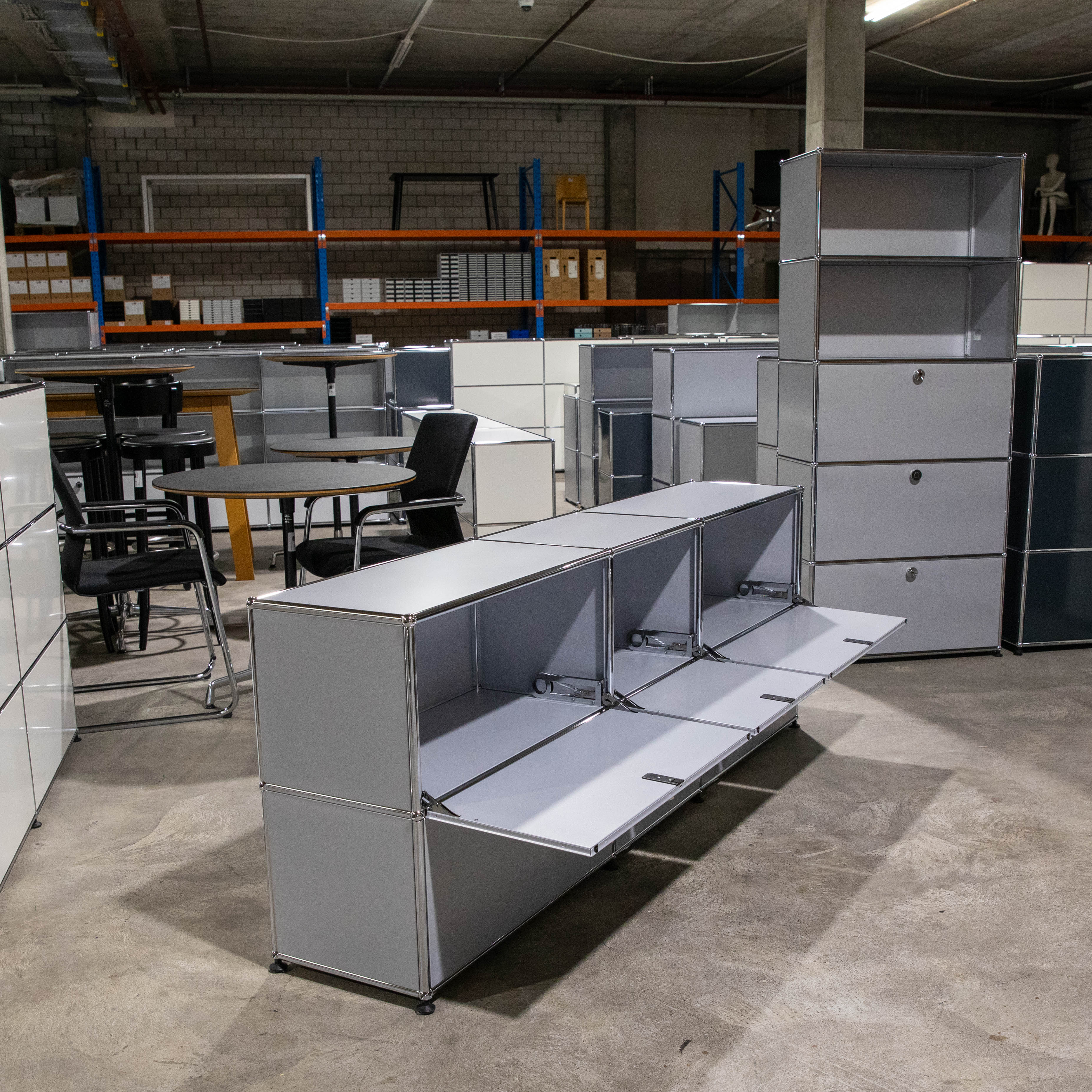 Ein elegantes USM Haller Sideboard in Mattsilber steht im Vordergrund, mit drei offenen Klapptüren, die leere Fächer zeigen. Teil weiterer modularer USM Möbel in Grau und Weiß, ausgestellt in einem hellen Lagerraum.