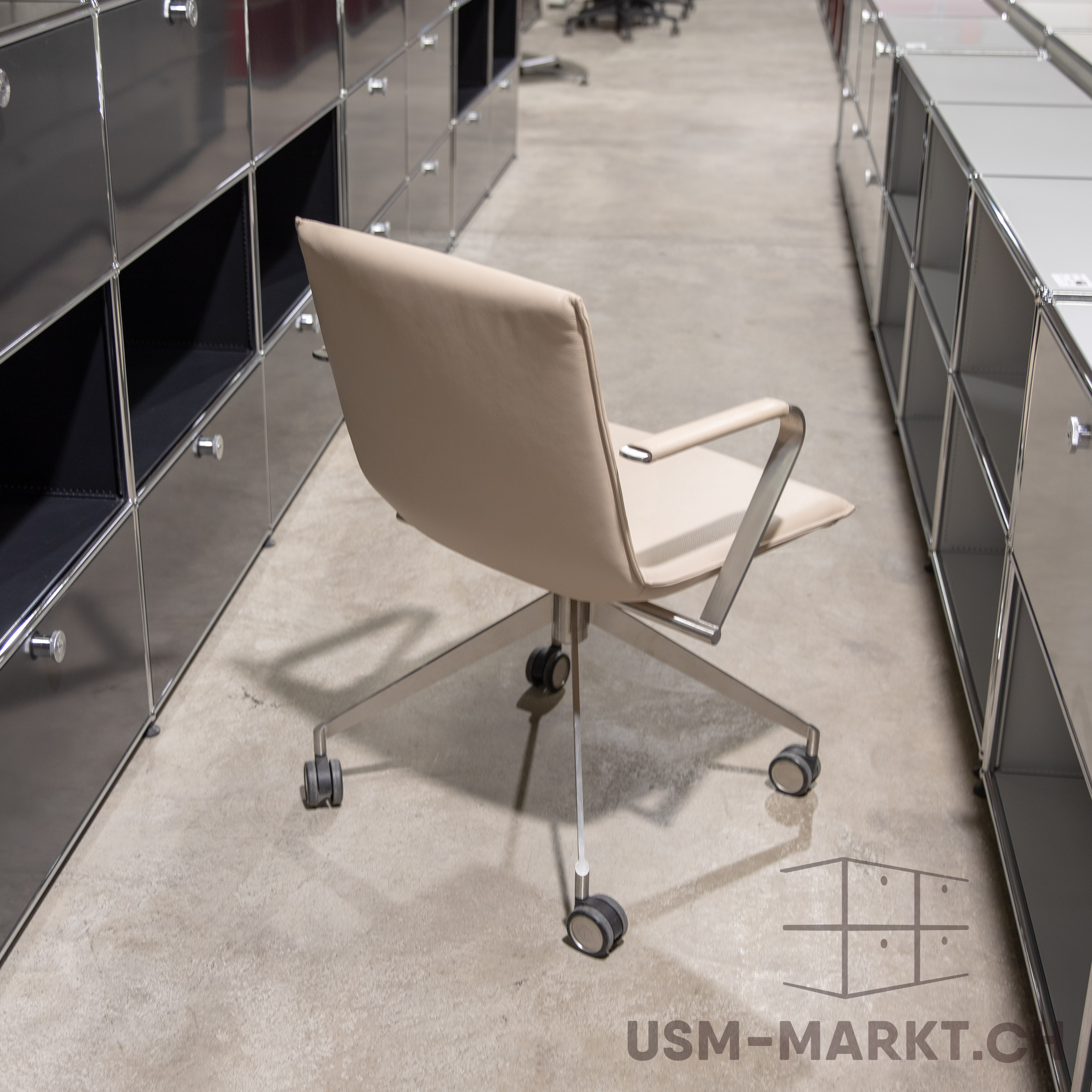 Beige office chair with upholstered backrest and seat, chrome base and castors on a light grey concrete floor. Facing away from the viewer in an aisle between two rows of modular dark shelving units with chrome frames.