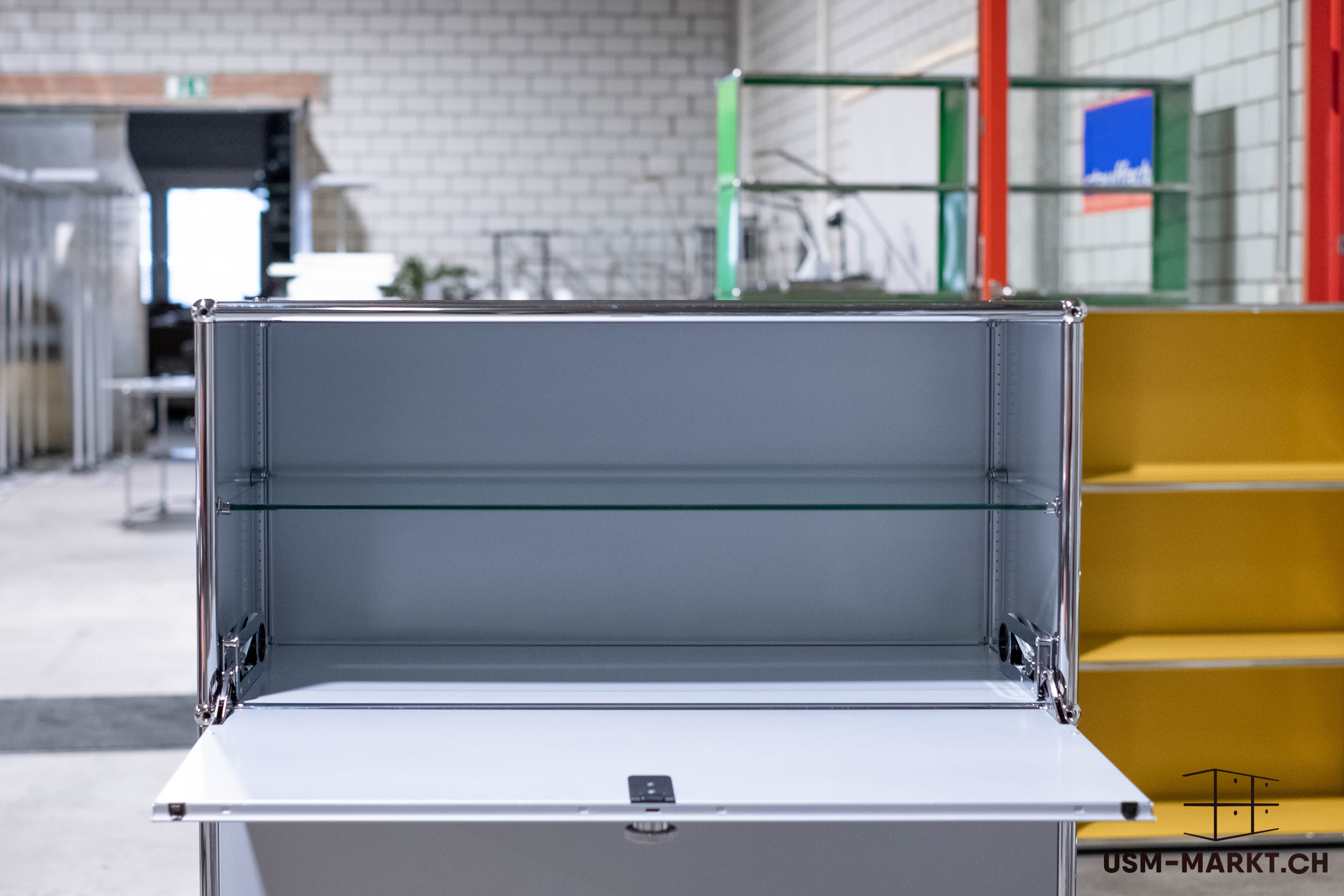 Open light grey modular cupboard with chrome detailing. The front flap opens downwards; inside is a glass shelf. In the background, out-of-focus shelves in a storeroom.