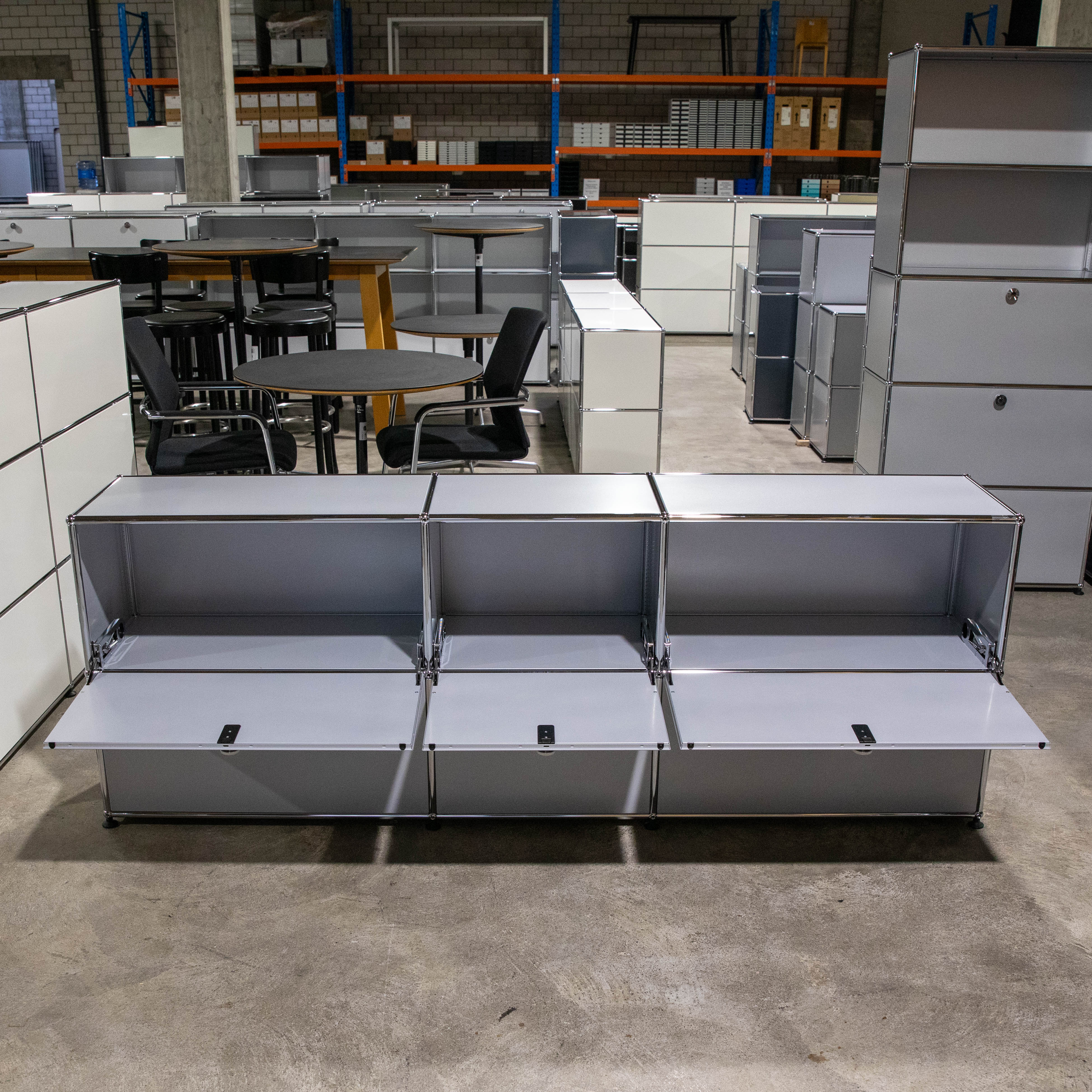 Vorderansicht eines gebrauchten USM Haller Sideboards in Mattsilber mit drei geöffneten Klapptüren, die den Blick ins leere Innere der Fächer freigeben. Modulare Konstruktion aus Chromrohren und Metallplatten in großer Halle.