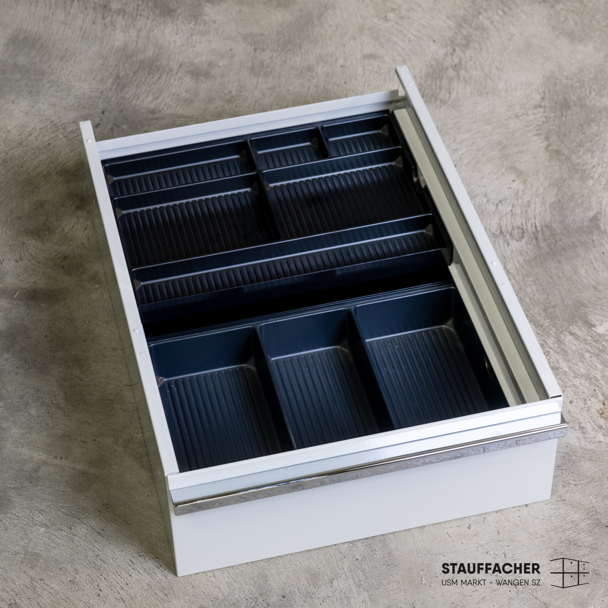 Mobile a cassetti aperto grigio chiaro con inserto organizzatore scuro a doghe. L’organizzatore ha scomparti di varie dimensioni per riporre gli oggetti in ordine. Il mobile poggia su base in cemento grigio, moderno e funzionale.
