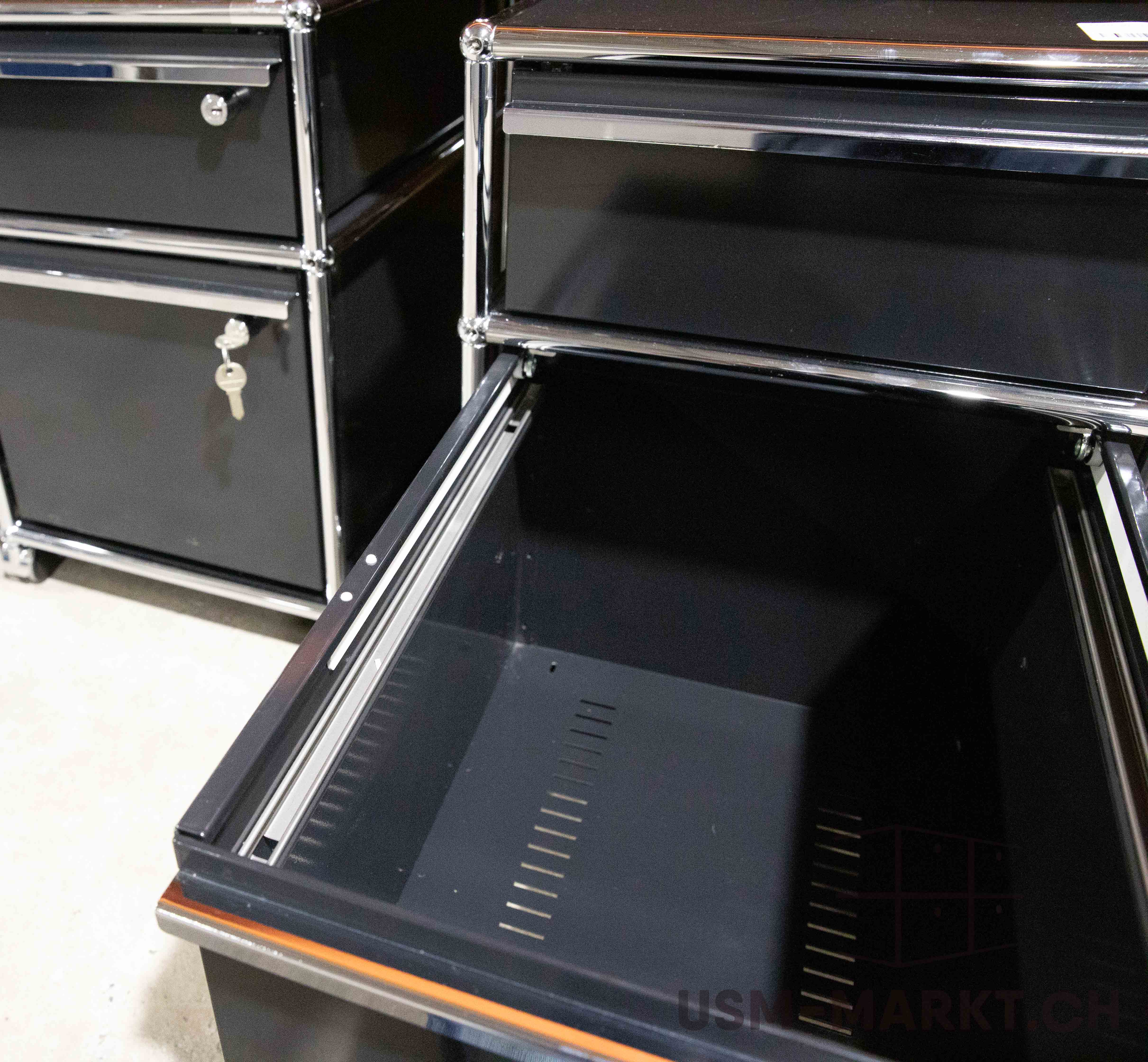 Offener, leerer Schubkasten eines schwarzen modularen Schranks mit glänzendem Chromrahmen; dunkles Inneres und seitliche Lochbleche sichtbar. Im Hintergrund weitere geschlossene Schubladen, eine mit Schlüssel im Schloss, auf hellem Boden.