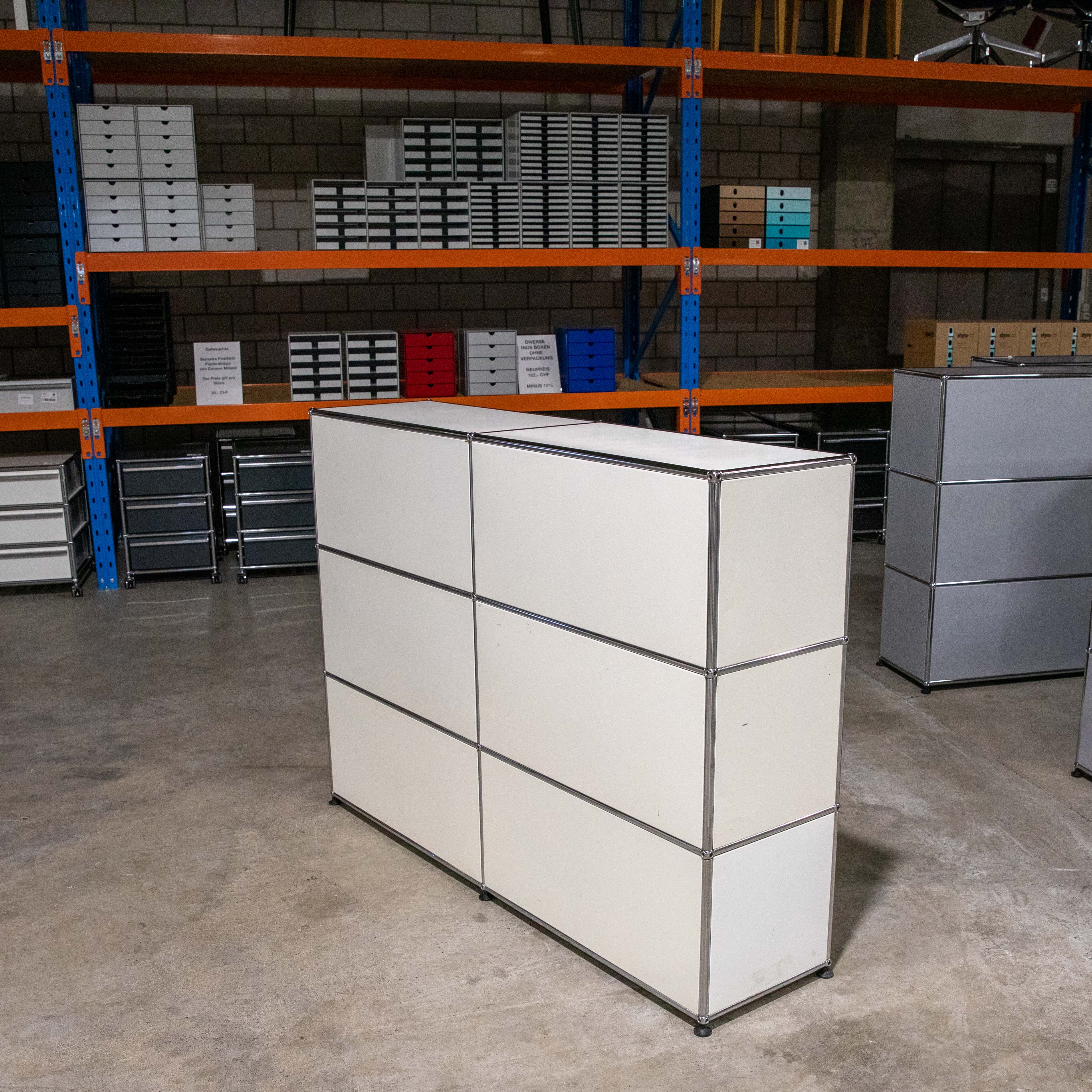 Großer cremefarbener modularer Schrank mit sechs Fächern und silbernen Rahmen auf Betonboden in Lagerraum. Hinten orange Regale mit blauen Stützen, gefüllt mit Büroablagen und Boxen in Weiß, Schwarz und Farben.