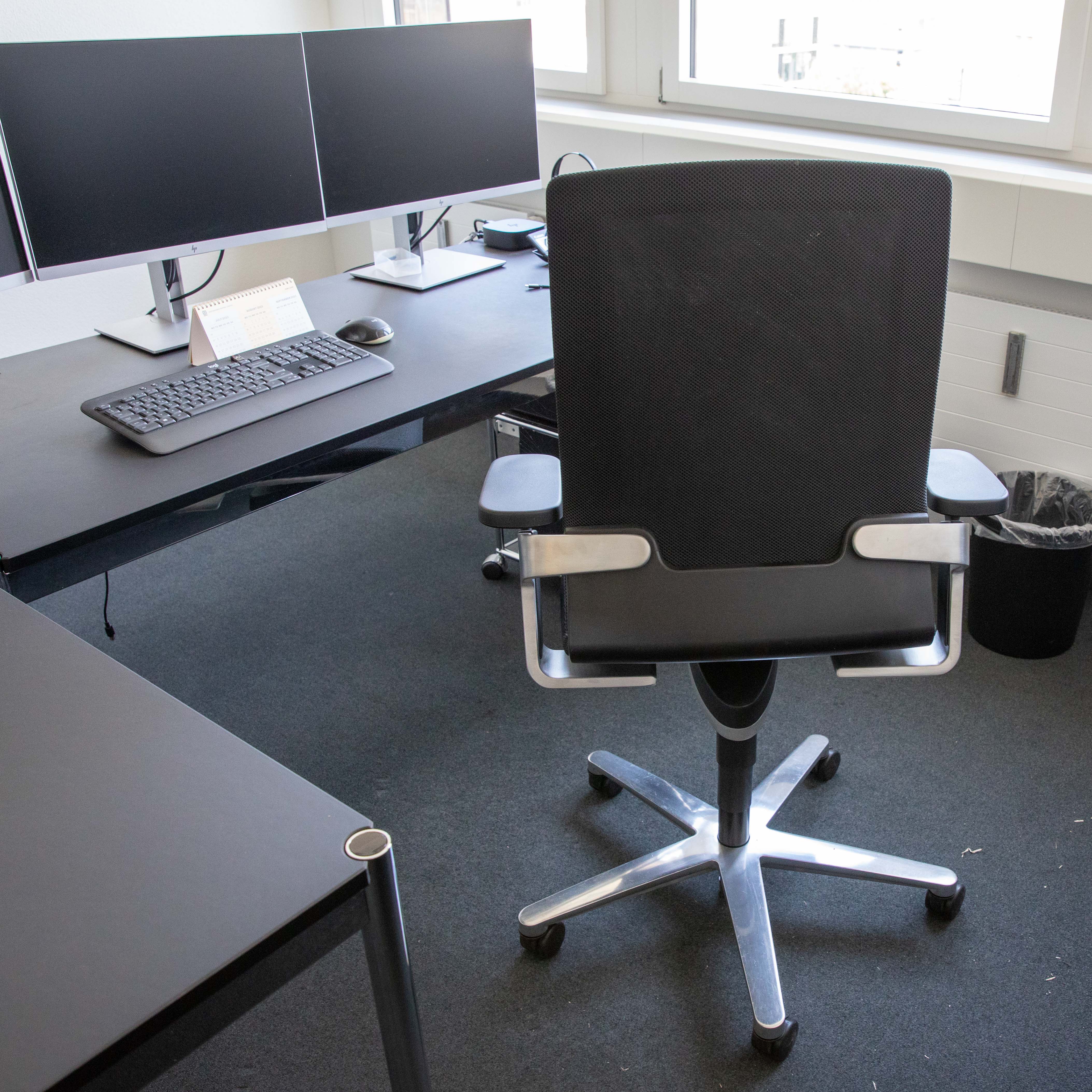 Schwarzer Wilkhahn ON Bürodrehstuhl mit Netzrückenlehne und Metallfußkreuz, von hinten. Vor dunklem Schreibtisch mit zwei Monitoren, Tastatur, Maus und Kalender. Im Hintergrund ein Fenster. Stuhl/Armlehnen mit Gebrauchsspuren.