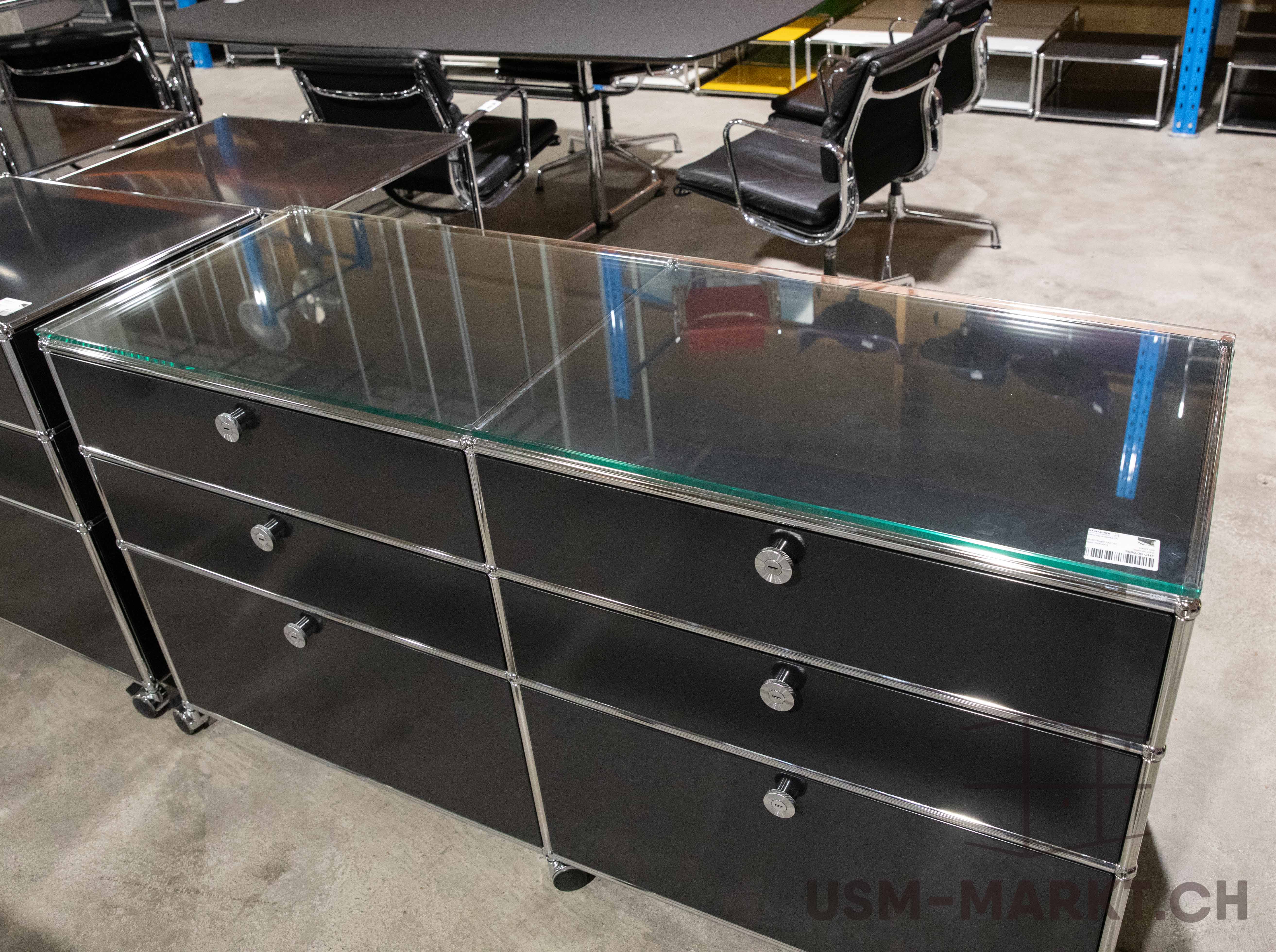 Two black, modular sideboards on castors with glossy glass tops and chrome-plated handles and frames. Each has three drawers with round metal locks. In the background, black office chairs and a large conference table. Concrete floor.