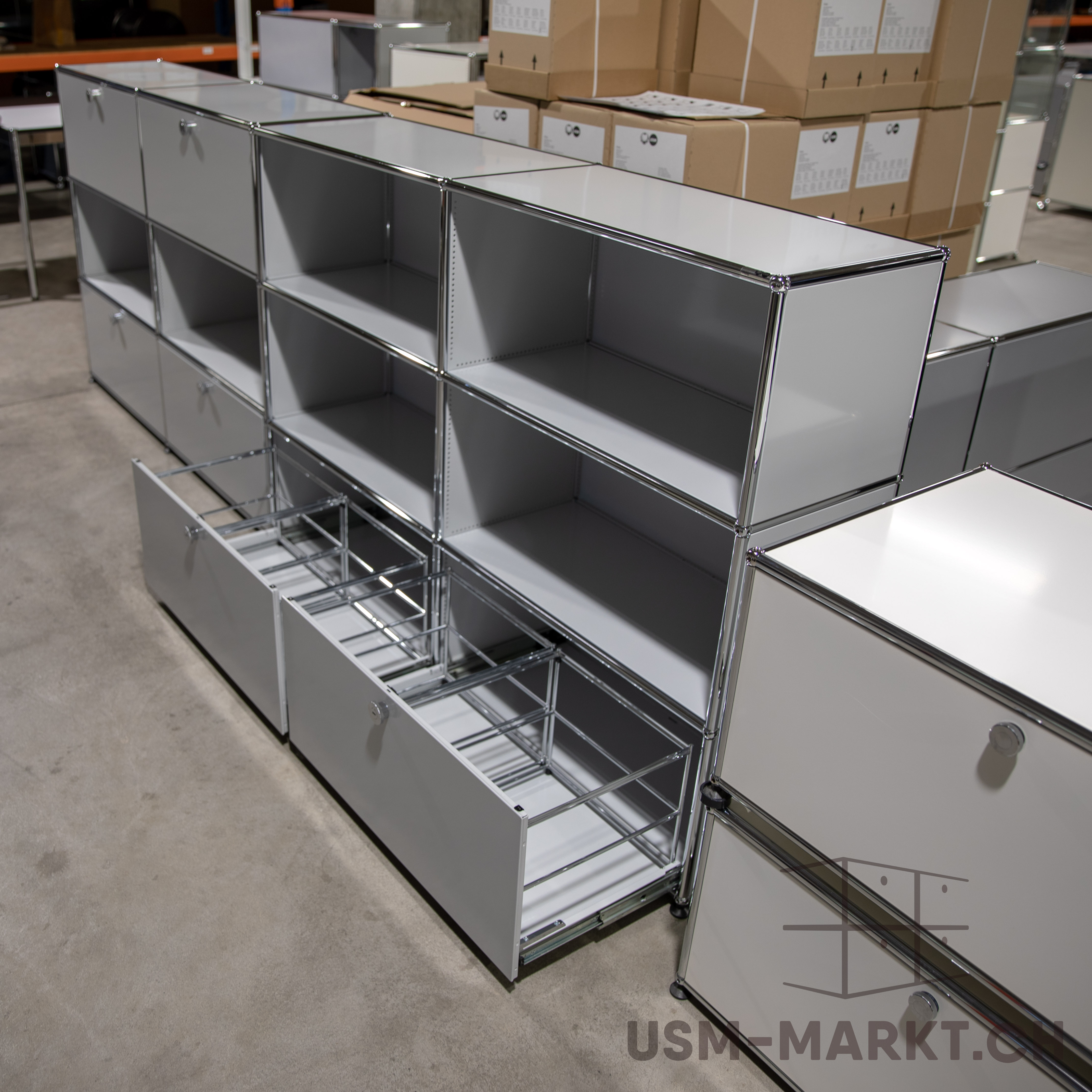 Mobili modulari USM Haller grigio chiaro su pavimento in cemento, con ripiani aperti e ante chiuse. Due cassetti aperti frontali mostrano telai metallici per raccoglitori sospesi. Sullo sfondo altri mobili e scatole.