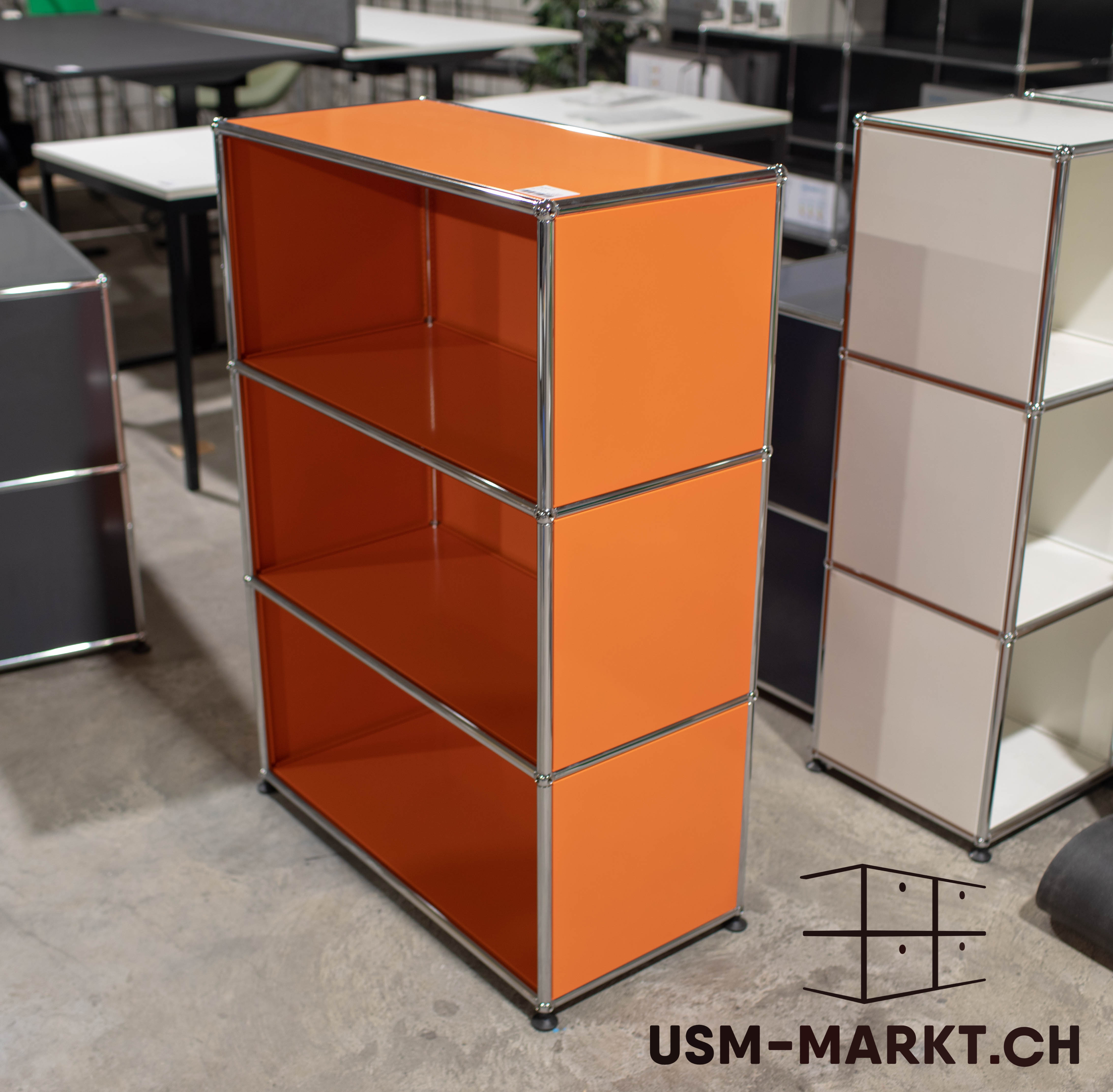 Una scaffalatura USM Haller arancione con tre ripiani aperti e barre cromate lucide è al centro su un pavimento in cemento grigio. In un ufficio moderno, altri mobili USM Haller e tavoli sfocati sullo sfondo.