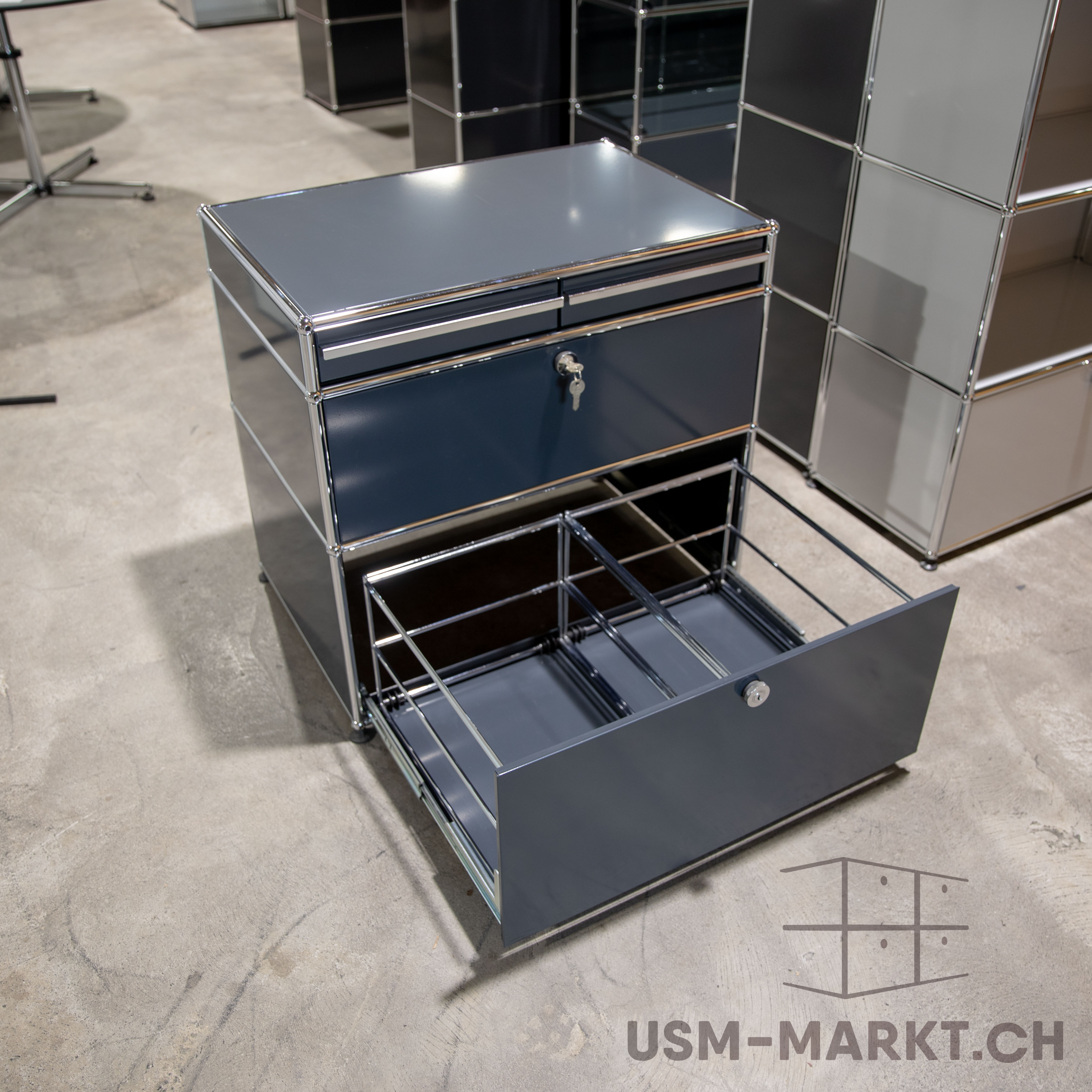 Dunkelgrauer USM Haller Rollcontainer mit drei Schubladen. Oben geschlossen mit Schlüssel. Unten ganz geöffnet, Metallgestell für Hängeregister sichtbar. Steht auf Betonboden, im Hintergrund weitere USM Möbel.