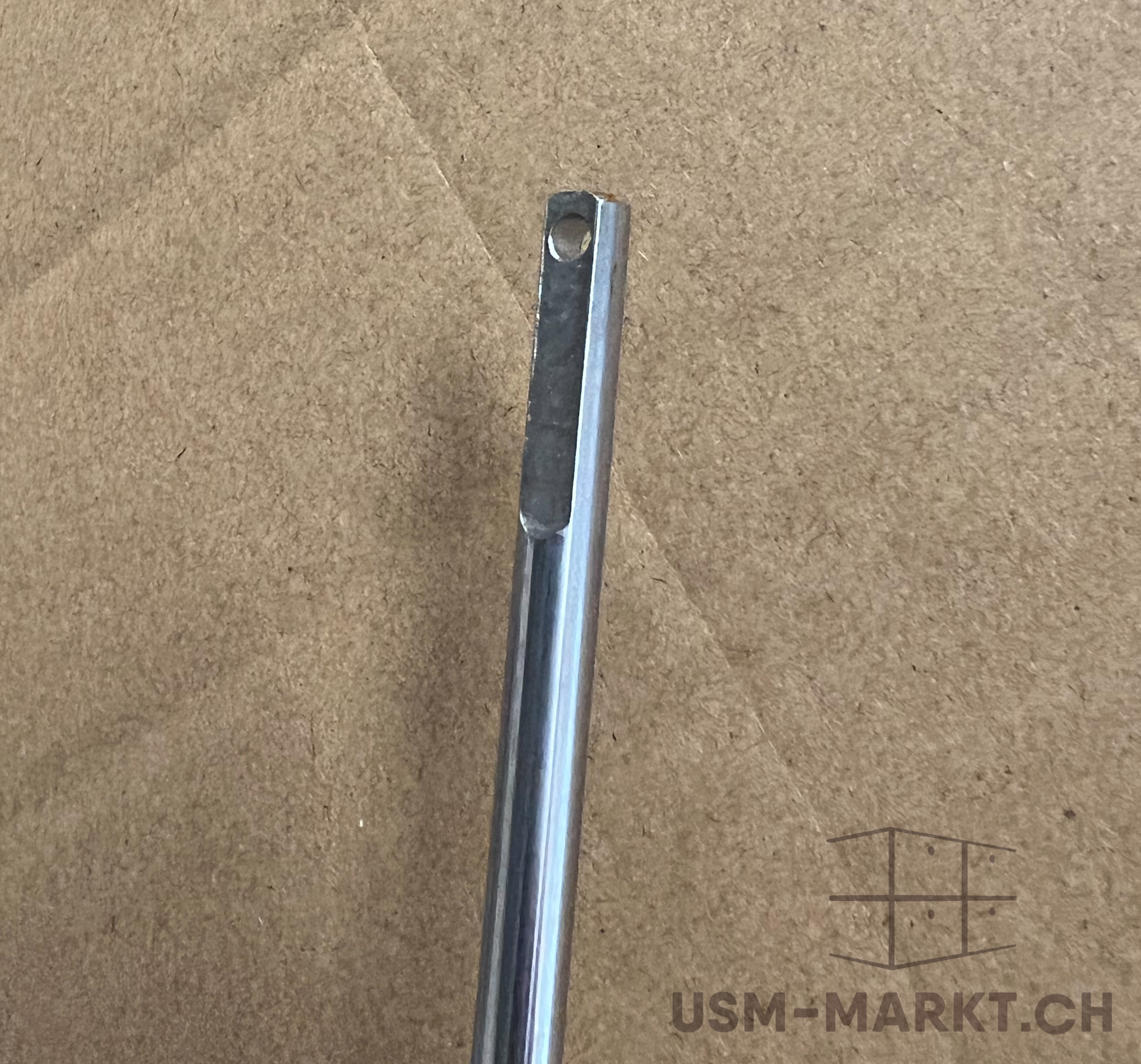 A shiny, metallic rod or pin is shown vertically against a textured, light brown background. The round body tapers into a flattened, rectangular upper section, featuring a small, circular hole.