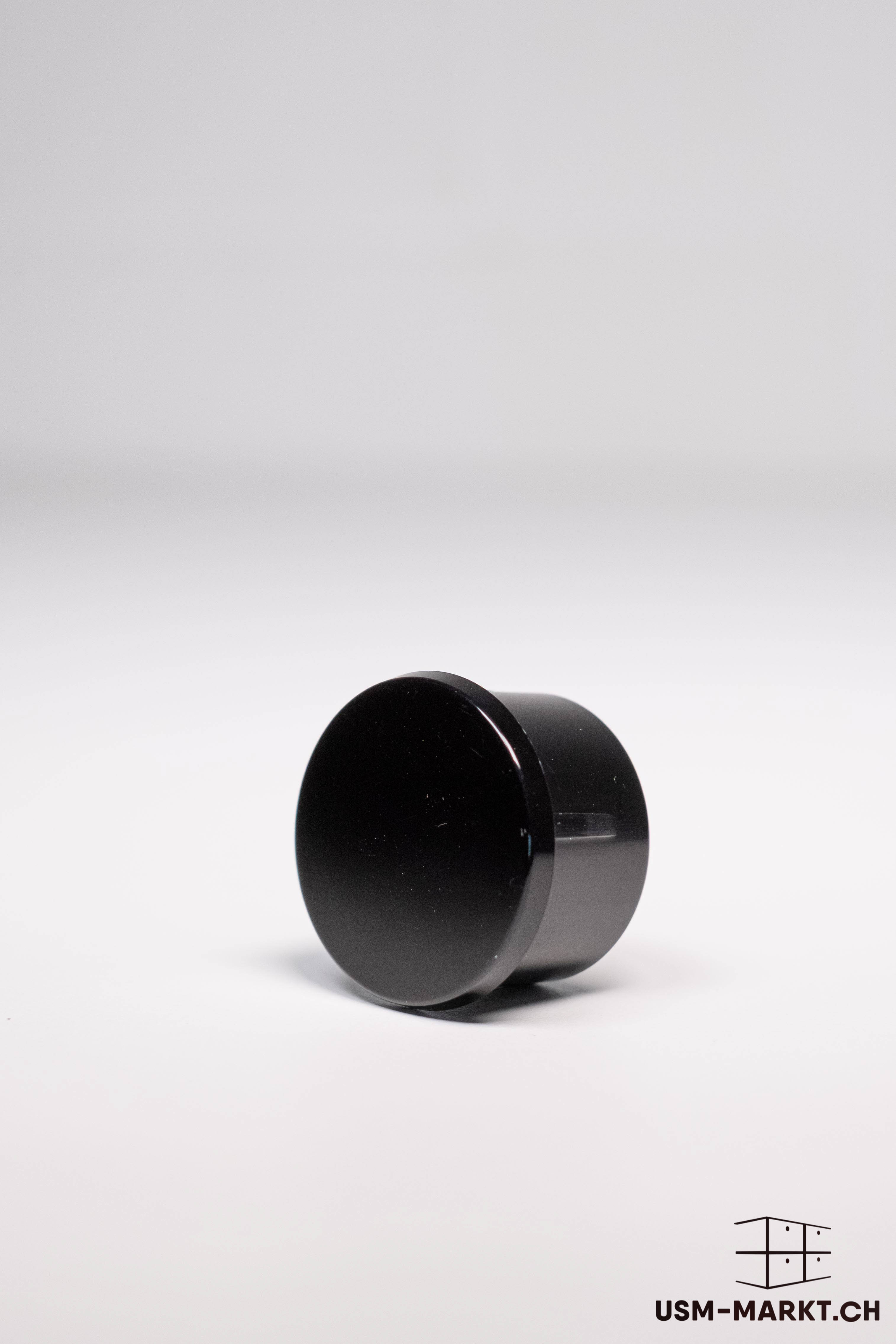 A glossy, cylindrical, black USM Haller connecting element lies horizontally on a light grey surface. The round end face is turned slightly towards the viewer. The surface reflects light and shows minimal irregularities.