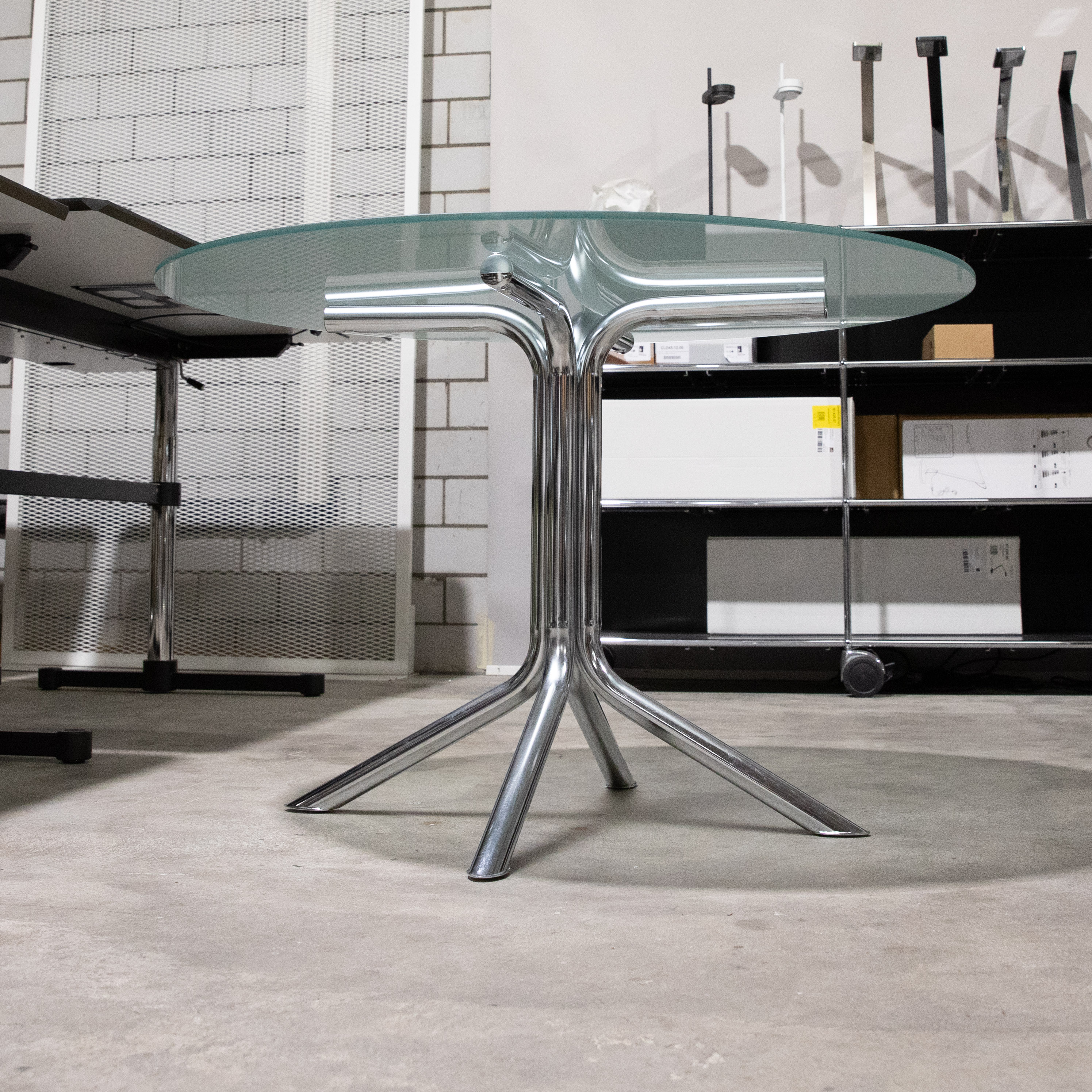 Table ronde dotée d'un plateau en verre dépoli élégant et légèrement translucide. Piètement chromé brillant avec une colonne centrale qui se divise en quatre pieds incurvés, design moderne. Posée sur un sol en béton gris clair dans la pièce.