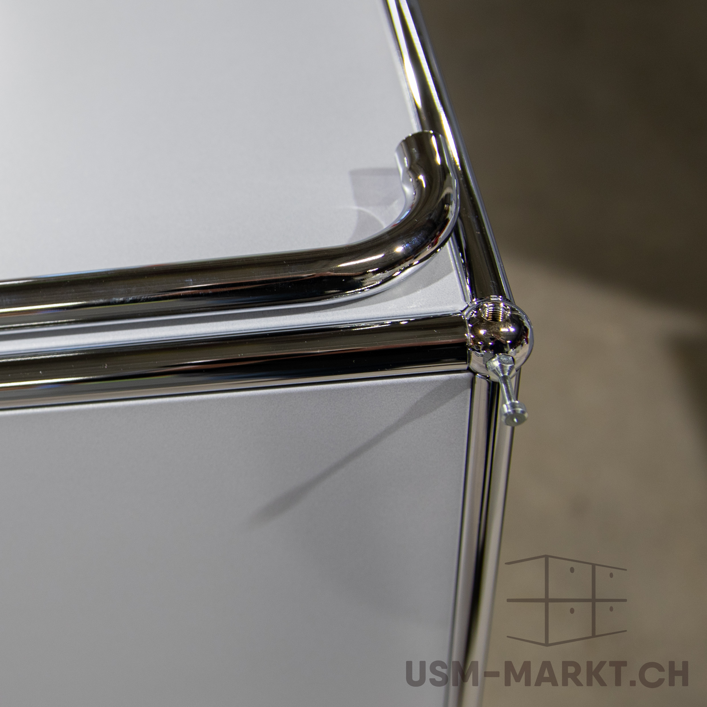 Nahaufnahme einer Möbelkante: verchromte Metallrohre an hellgrauer Platte, verbunden über ein kugelförmiges Gelenk mit sichtbarer Schraube. Minimalistisches Design, weiche Schatten.
