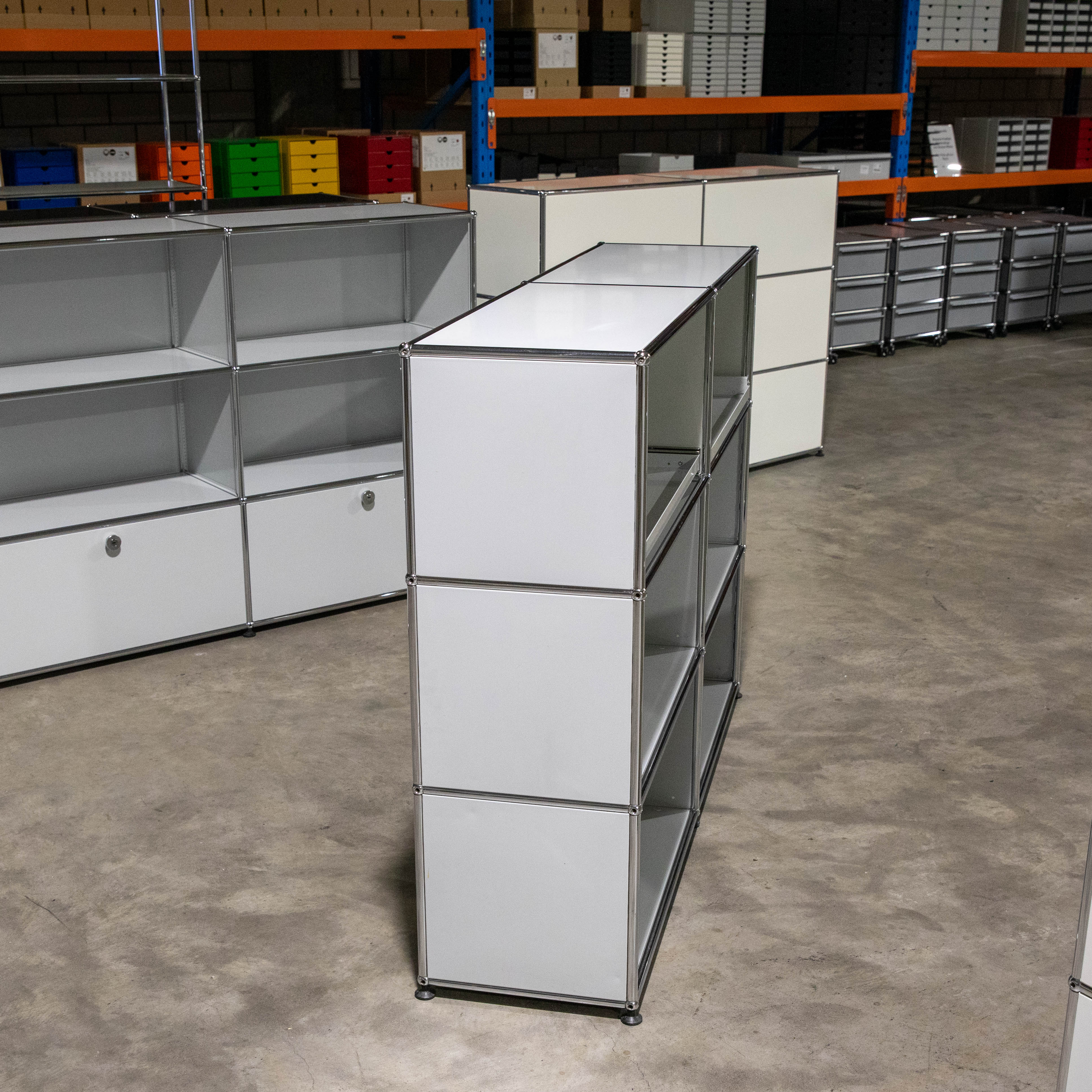 A narrow, light grey USM Haller shelving unit with three open compartments and closed side panels stands on a concrete floor in a storeroom. In the background, more USM Haller furniture and shelving are visible, holding colourful boxes and cartons.