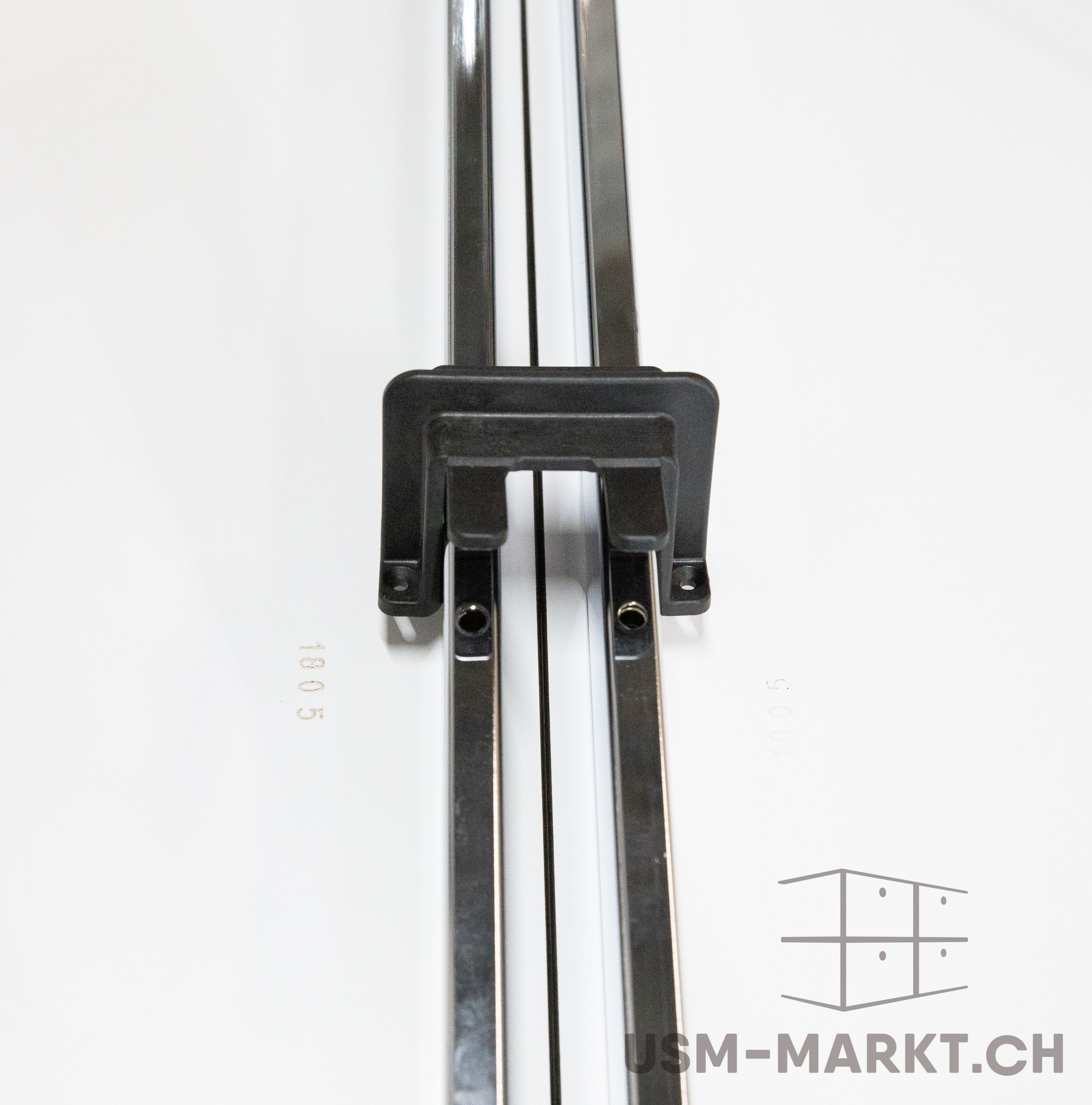 A black plastic element connects two parallel, polished metal rods mounted vertically on a white surface. It encircles the rods and has small round holes on the sides. The golden numbers ‘1805’ are printed on it.