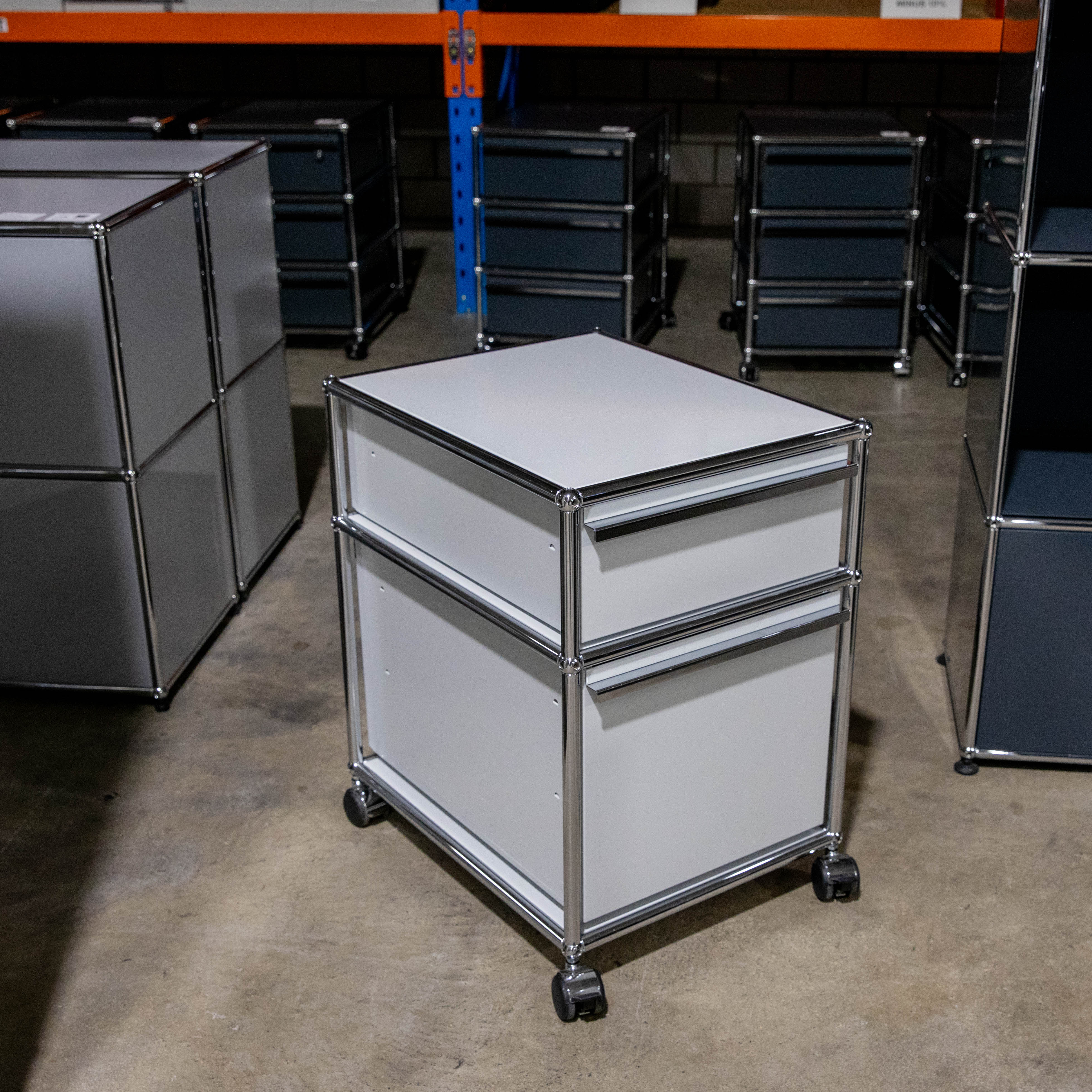 Ein hellgrauer, modularer Rollcontainer mit zwei Schubladen und poliertem Chromrahmen steht auf Rollen im Vordergrund einer Lagerhalle. Im Hintergrund sind weitere dunkle Schranksysteme und orange-blaue Lagerregale auf einem Betonboden sichtbar.