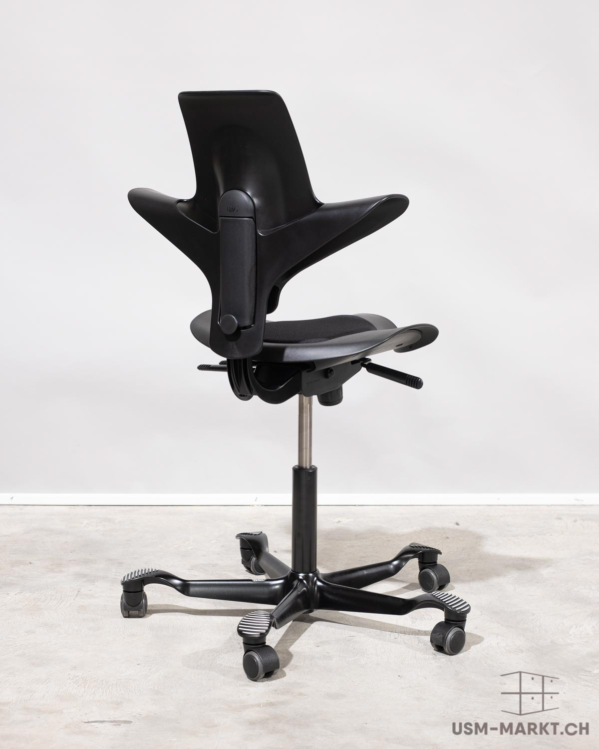 Sedia da ufficio ergonomica nera moderna, vista leggermente di profilo da dietro, davanti a parete grigio chiaro e pavimento in cemento. Schienale curvo con braccioli integrati, seduta imbottita, base a cinque razze con rotelle. Al centro.