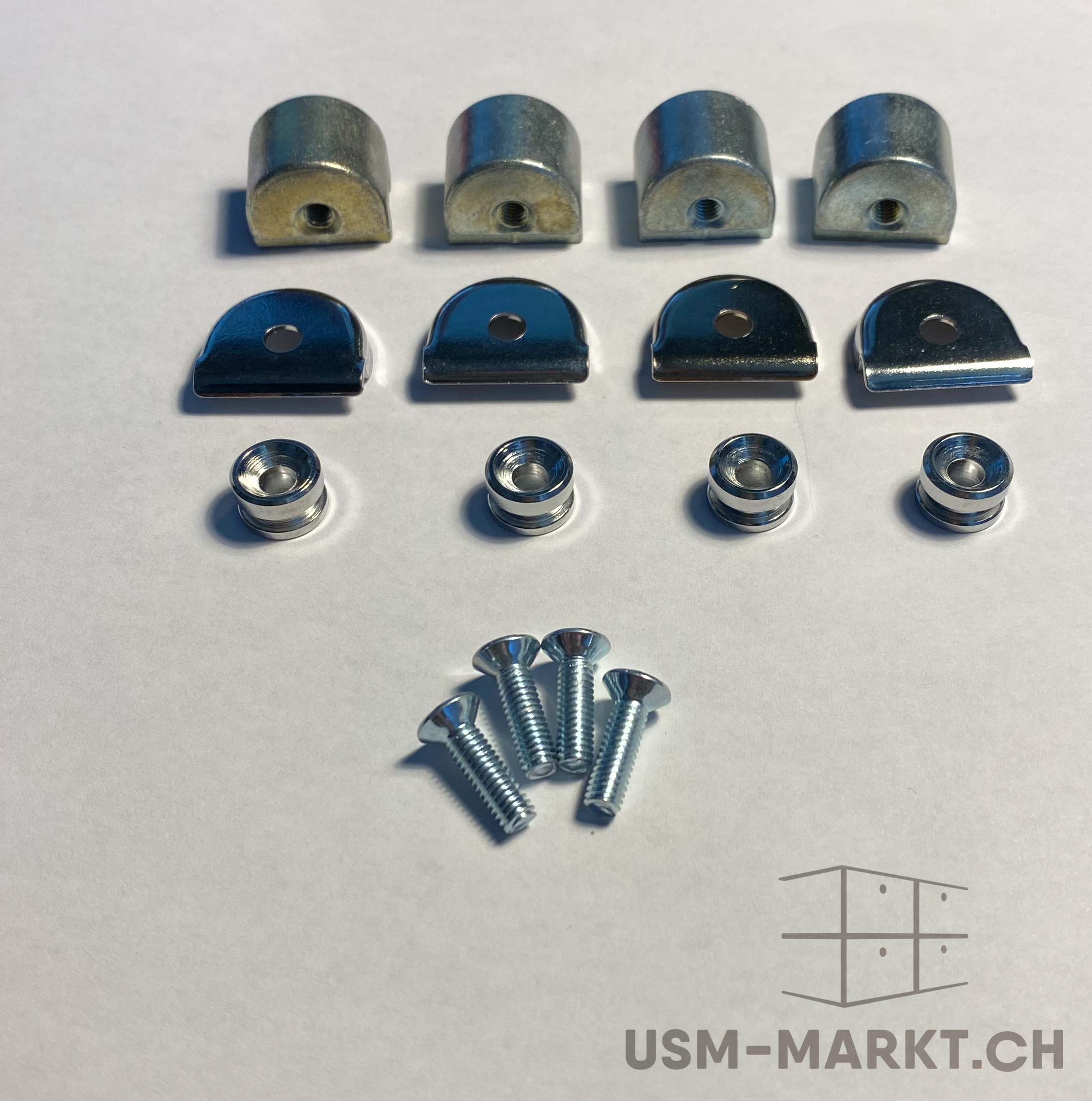 Accessori metallici per mobili ed elementi di fissaggio su sfondo bianco: in alto quattro pattini semicilindrici con fori filettati, sotto quattro lamiere lucide e ricurve, quattro bussole e quattro viti a testa svasata.